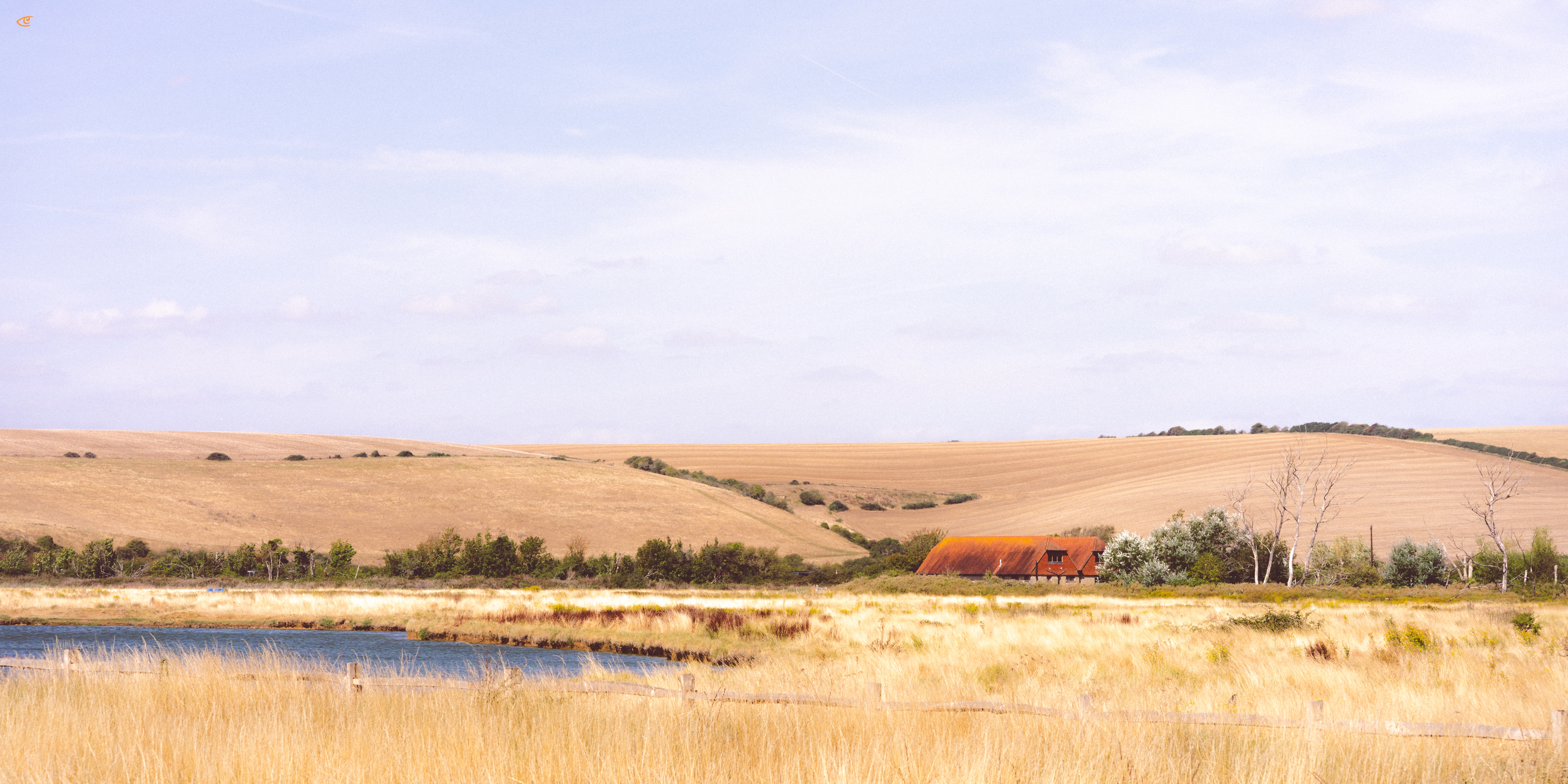 Wide rural landscape with a small pond in the foreground, tall dry grass and shrubs, rolling tan fields in the distance, and a red-roofed farmhouse near the horizon under a pale blue sky.
