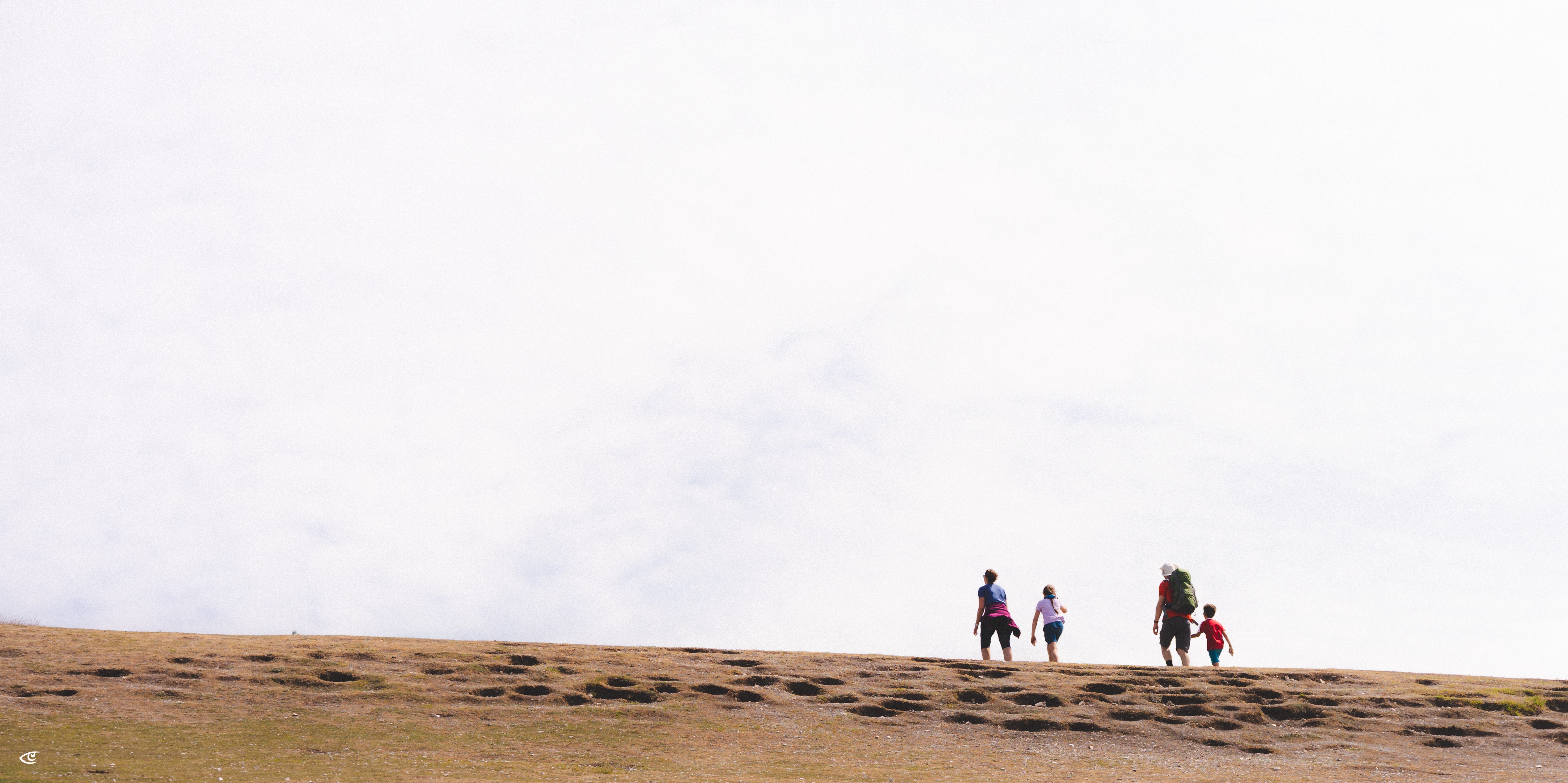 Four hikers walking along a grassy ridge line, silhouetted against a bright overcast sky, with open field in the foreground.