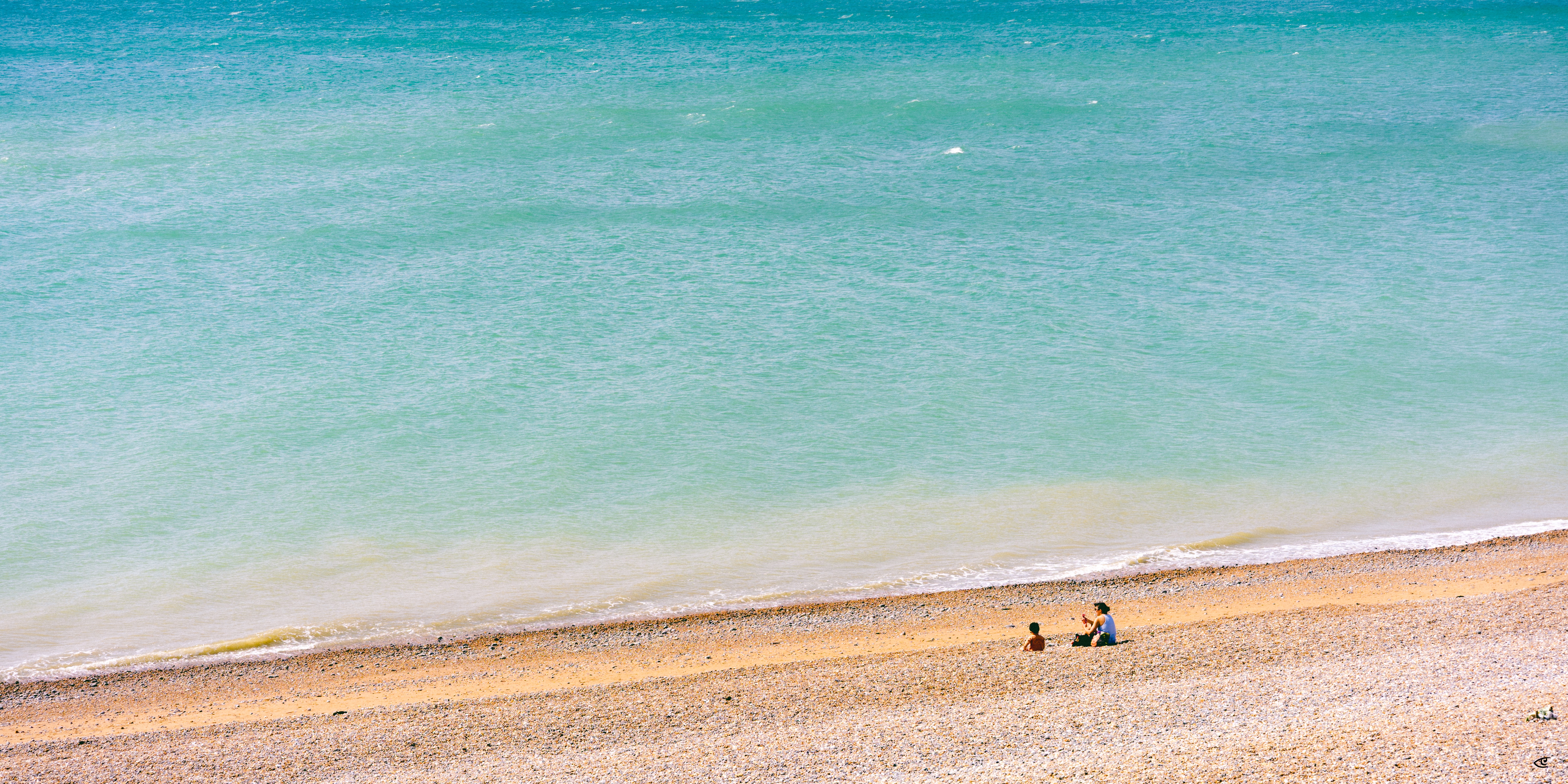 Aerial view of turquoise ocean meeting a narrow sandy beach, with two people sitting near the shoreline on the right.