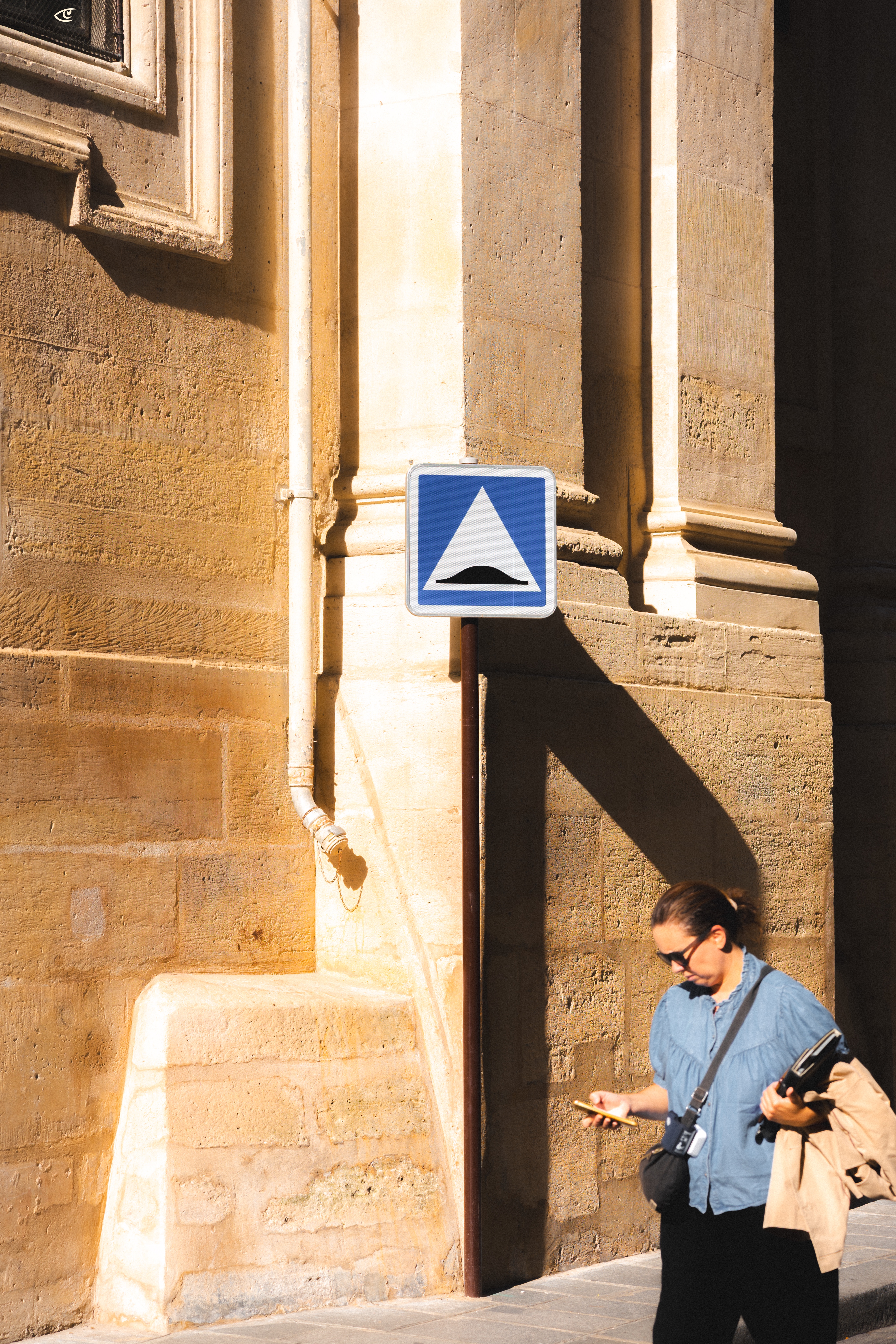 Blue and white road sign with a speed bump symbol on a pole against a sunlit stone building wall, casting a long shadow; woman in sunglasses walking at the bottom right while looking at her phone and carrying a jacket and bag.
