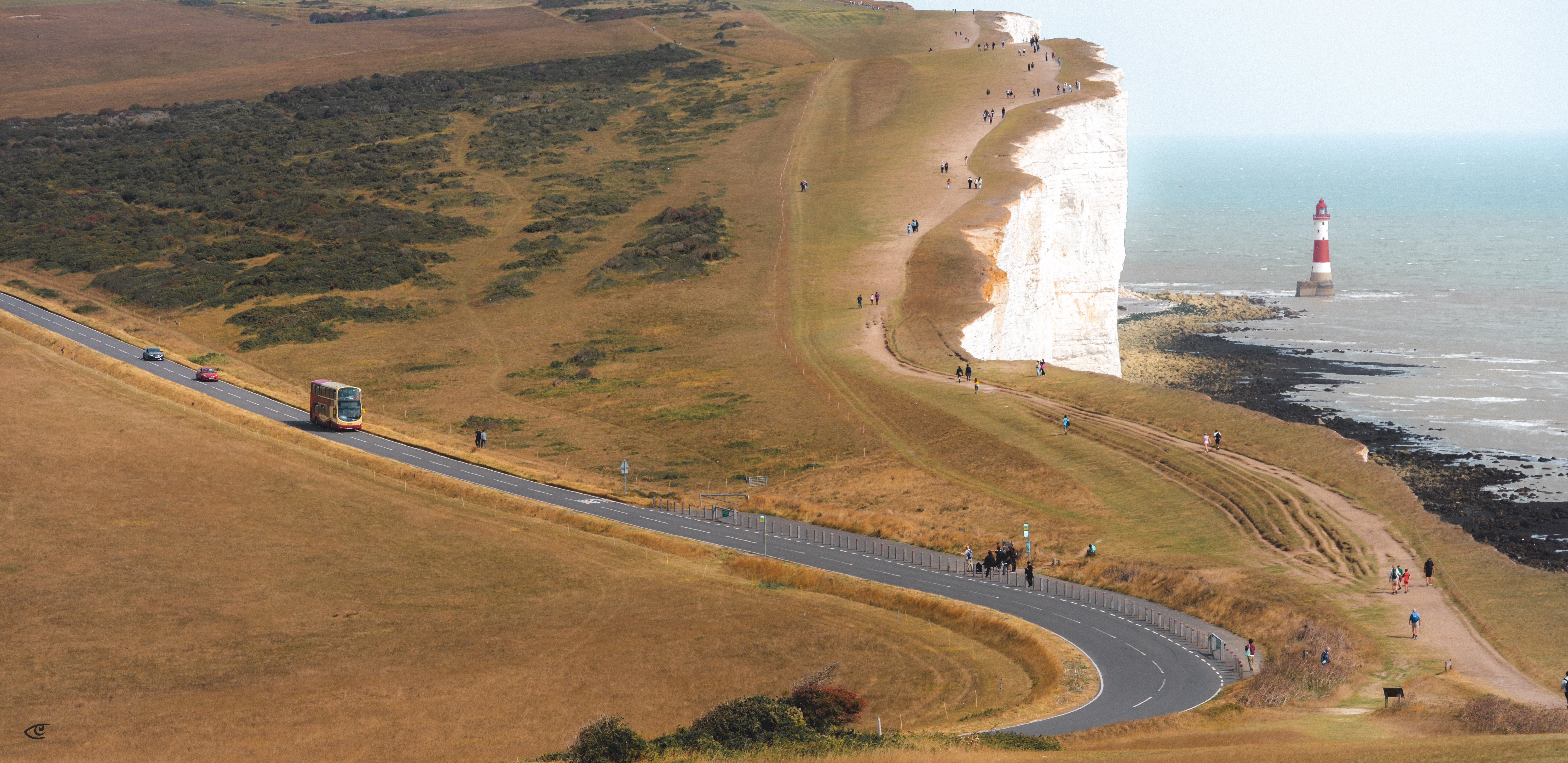 Aerial view of a coastal cliff and grassy headland with a winding road in the foreground, a bus and cars traveling along it, and small groups of people walking near the cliff edge; an inset photo at right shows a red-and-white striped lighthouse on rocks beside the sea.