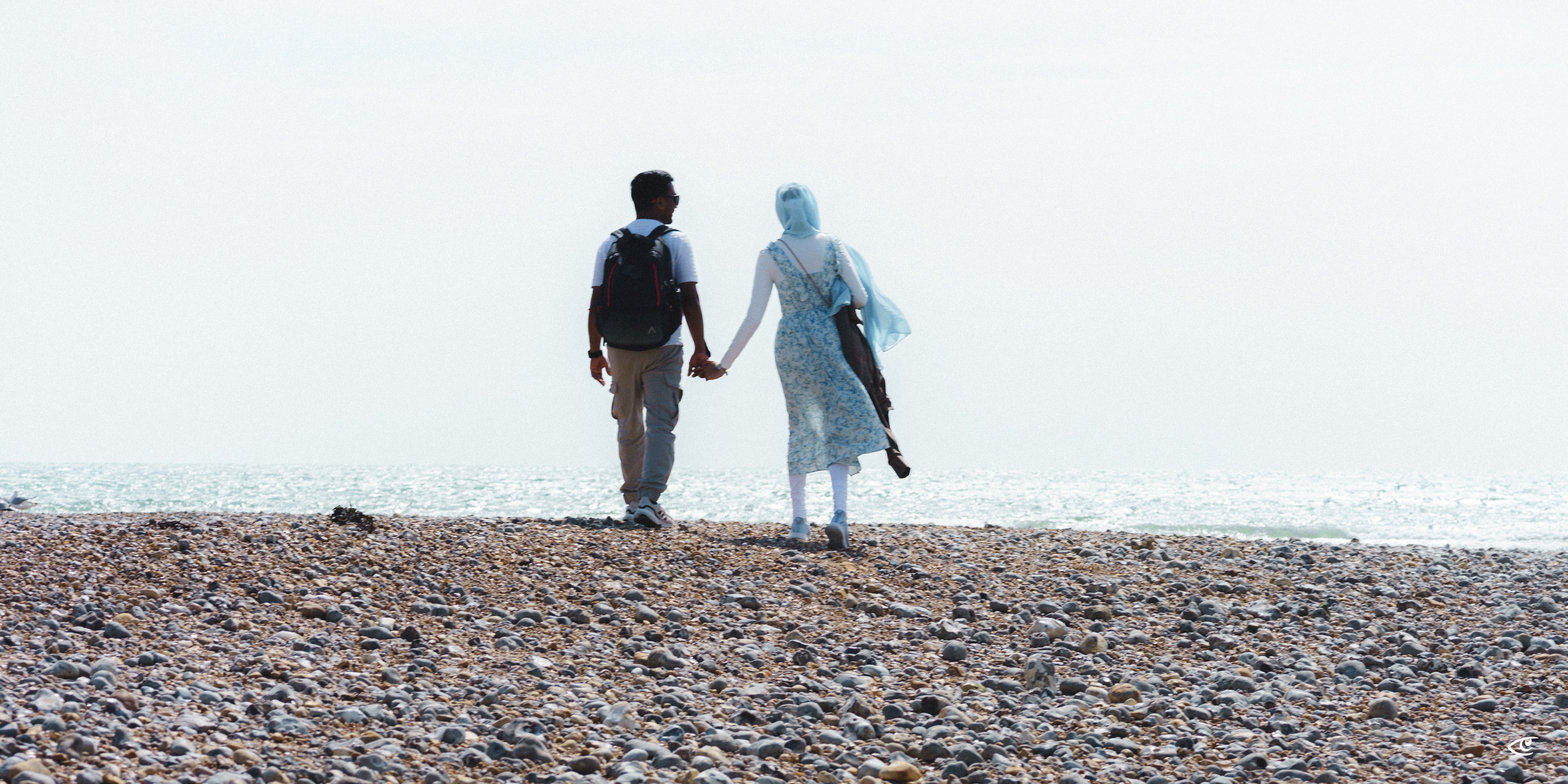 Two people holding hands on a rocky beach, seen from behind, facing the ocean horizon; one wears a backpack and the other wears a light blue headscarf and carries shoes.