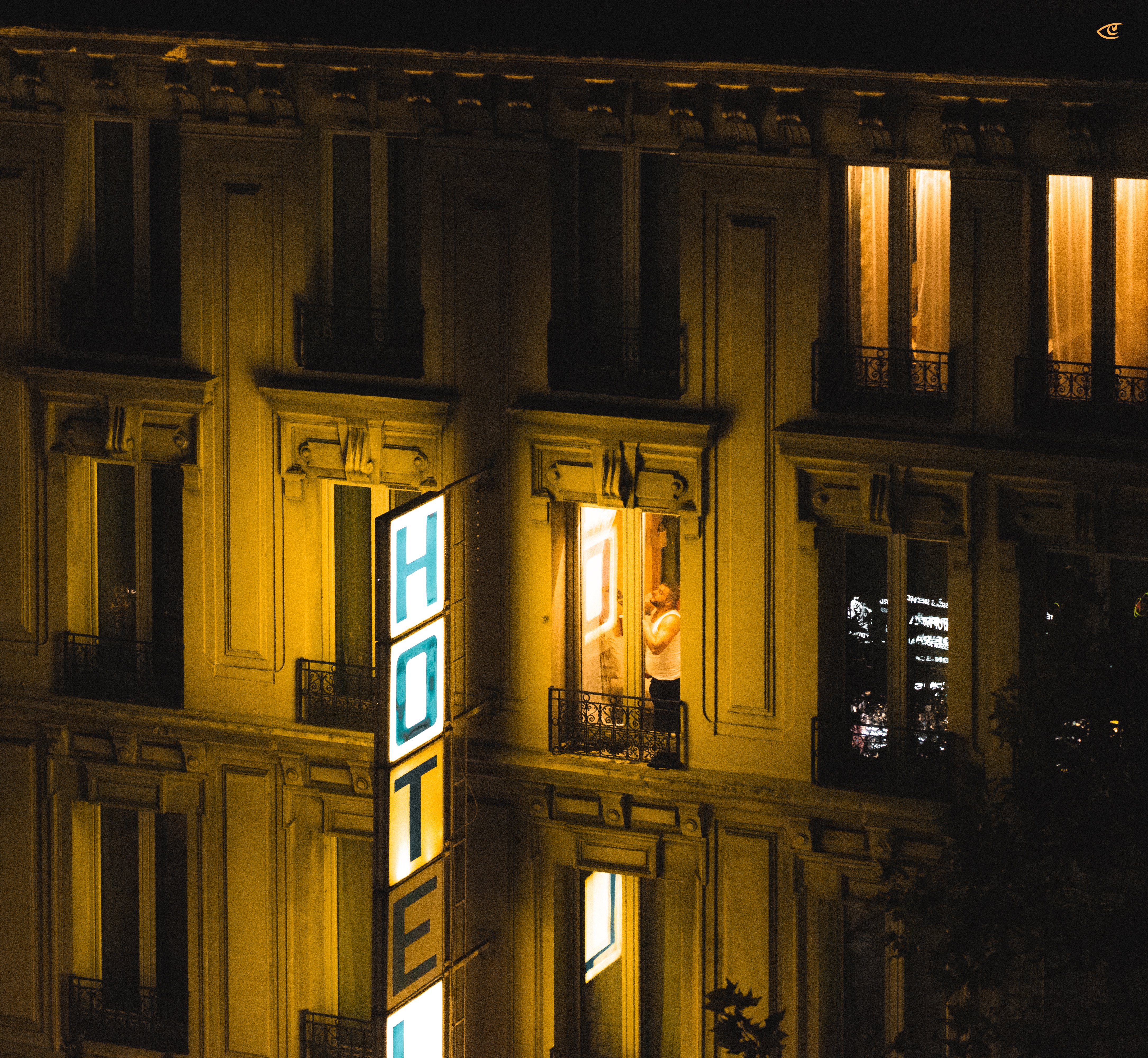 Nighttime view of a multi-story building with a vertical illuminated “HOTEL” sign on the facade; several windows glow with warm light, including one open window where a person stands on a small balcony railing.
