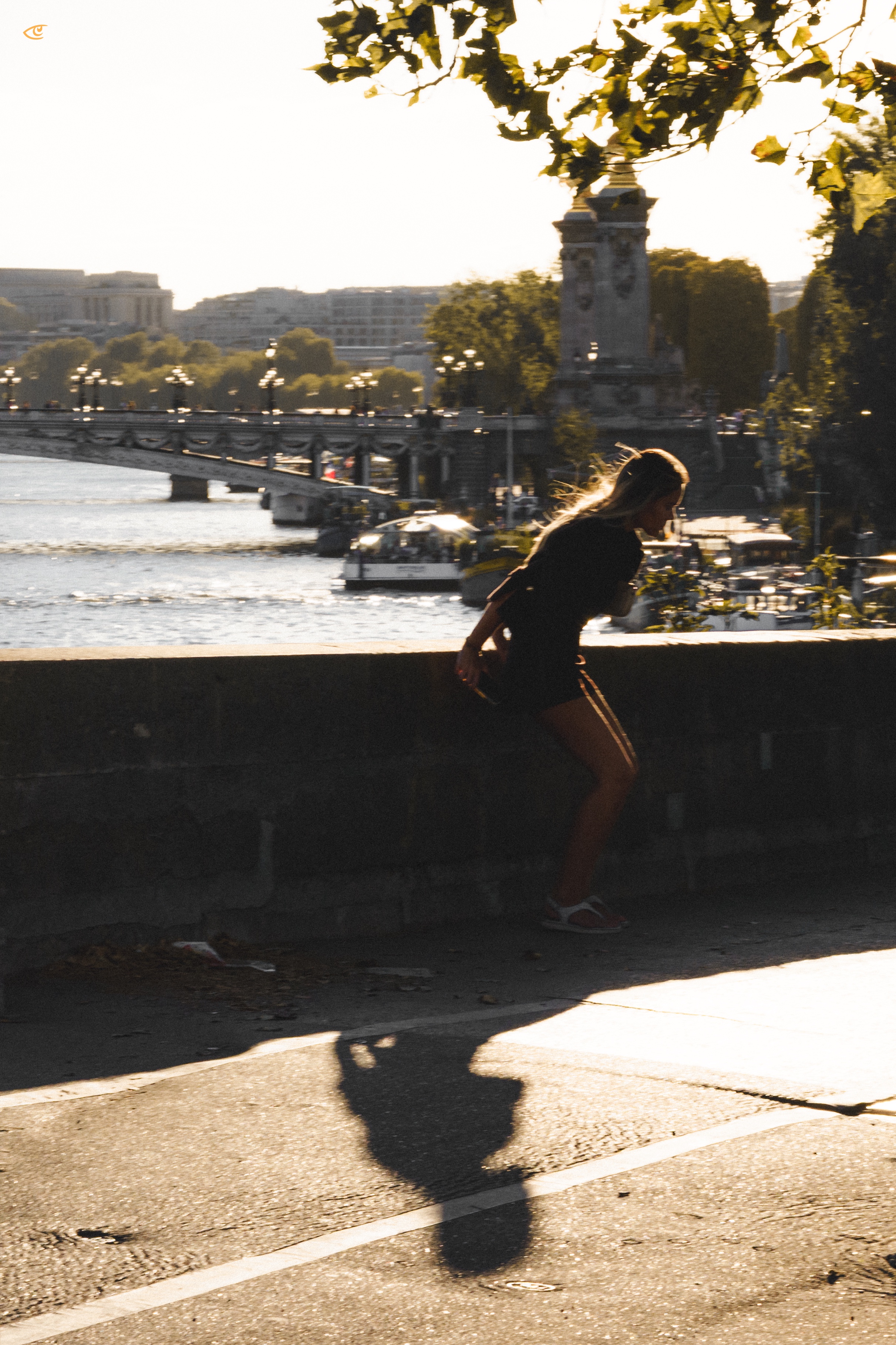Backlit silhouette of a woman leaning on a stone riverside wall, with her shadow stretching across the pavement; a bridge, riverboats, trees, and city buildings appear in the background, with leafy branches at the top of the frame.