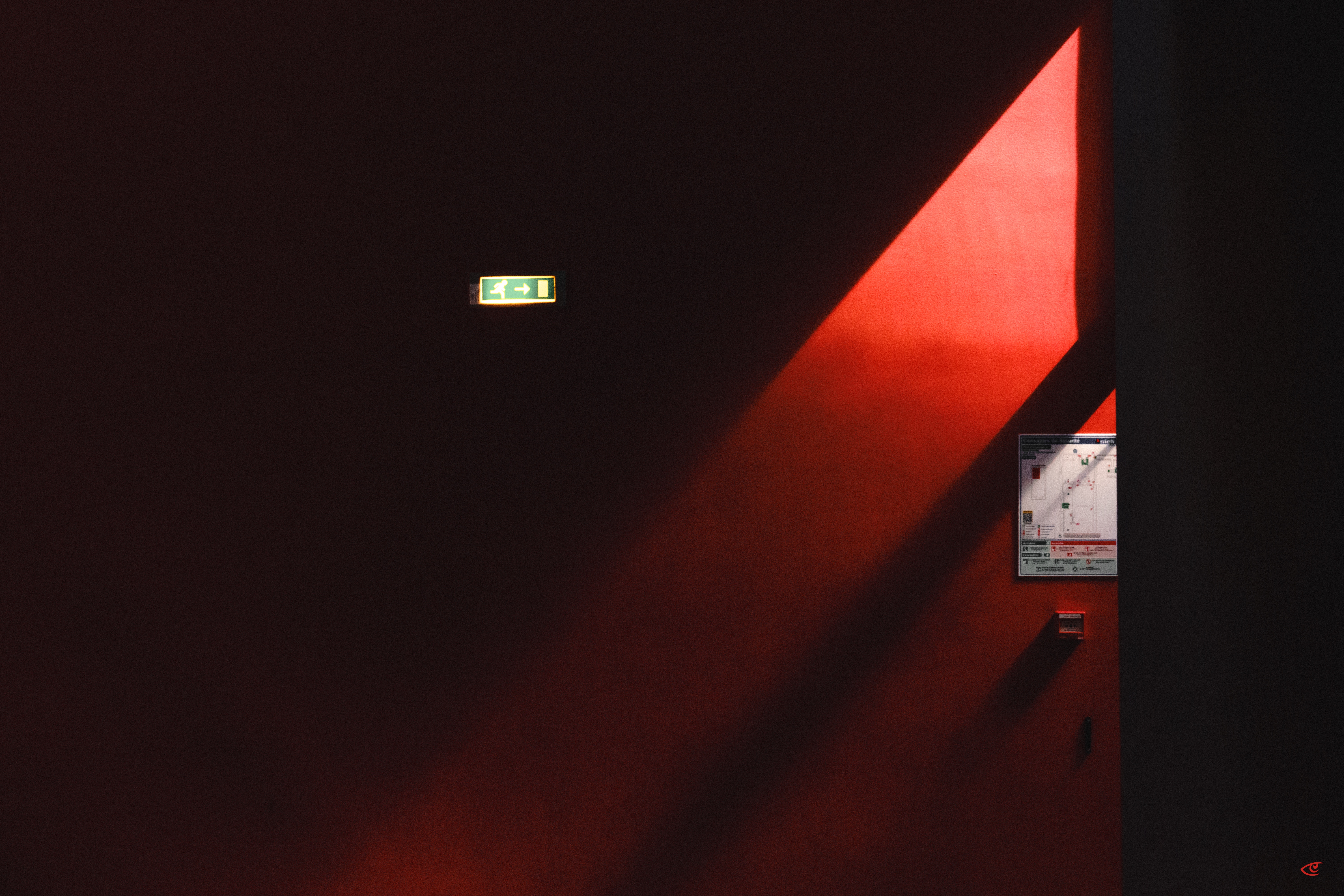 Dark interior wall with a diagonal beam of red light; a green illuminated exit sign near the upper center-left and a small wall-mounted panel on the right.