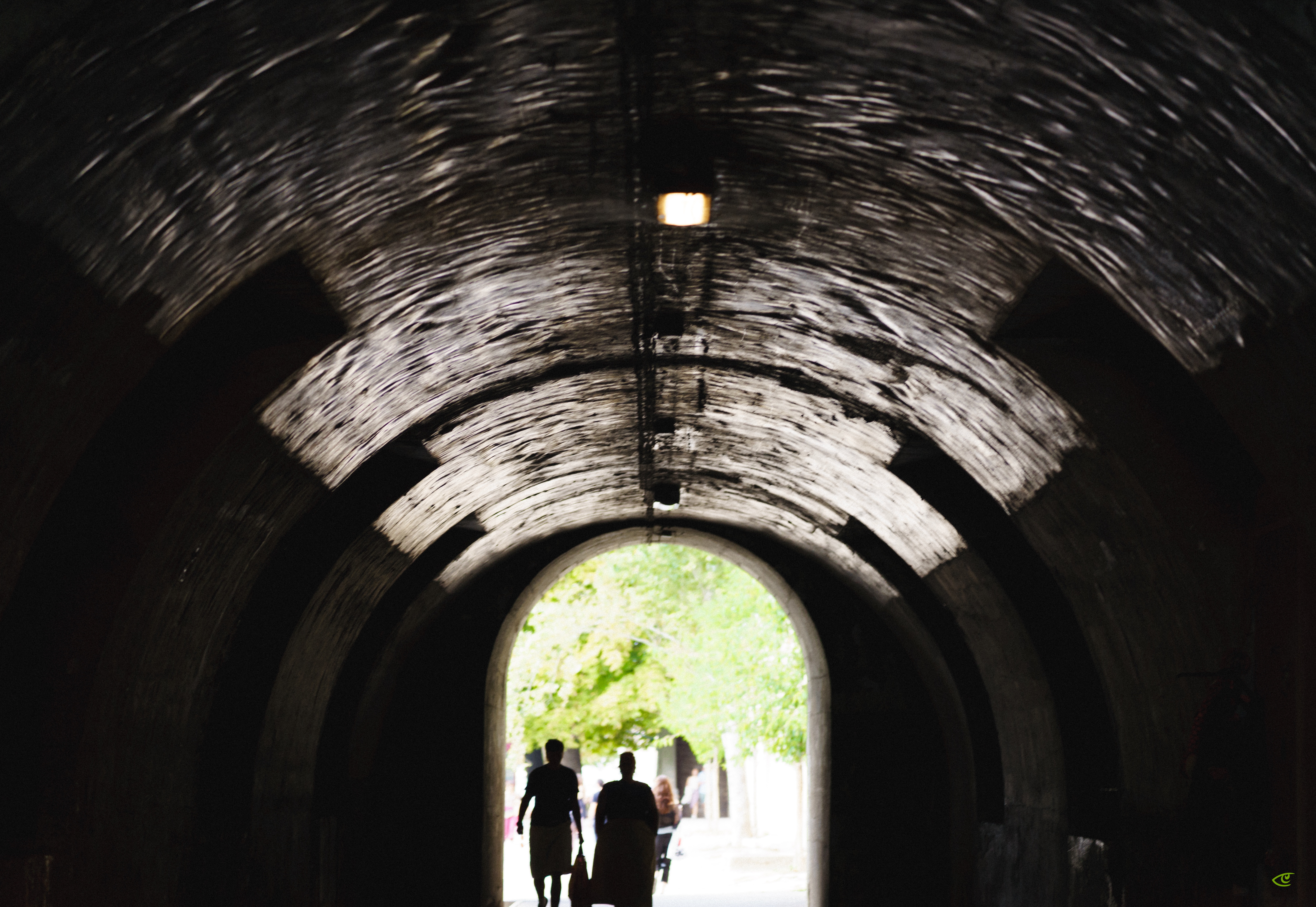 Silhouettes of two people walking through a dark, arched tunnel toward a bright exit with green trees visible outside; curved tunnel ceiling with a single light fixture overhead.