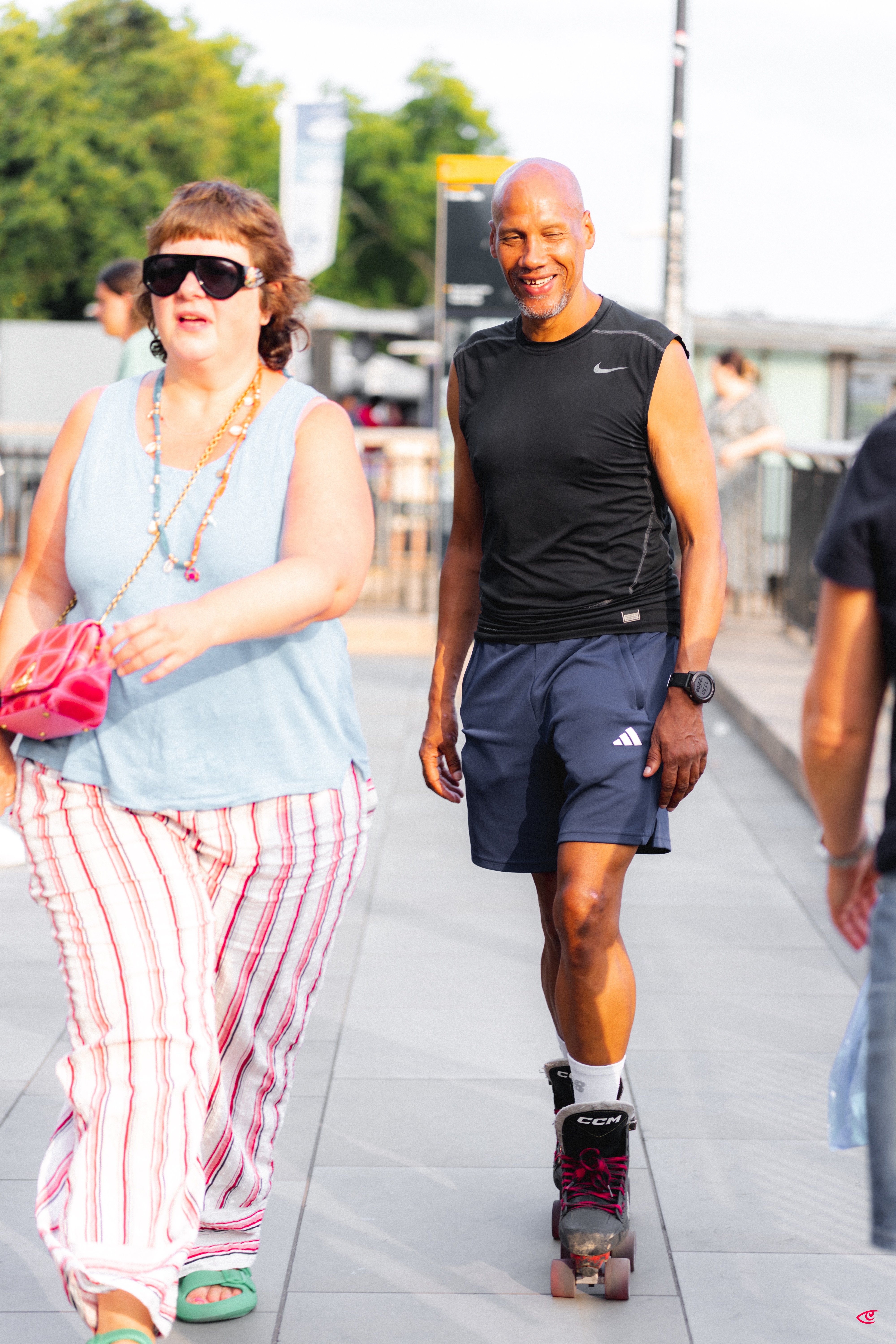 Two people on a city walkway: a woman in sunglasses wearing a light blue tank top and striped pants carrying a pink purse in the foreground, and a man in a black sleeveless shirt and navy shorts rollerblading behind her; trees, railings, and other pedestrians in the background.