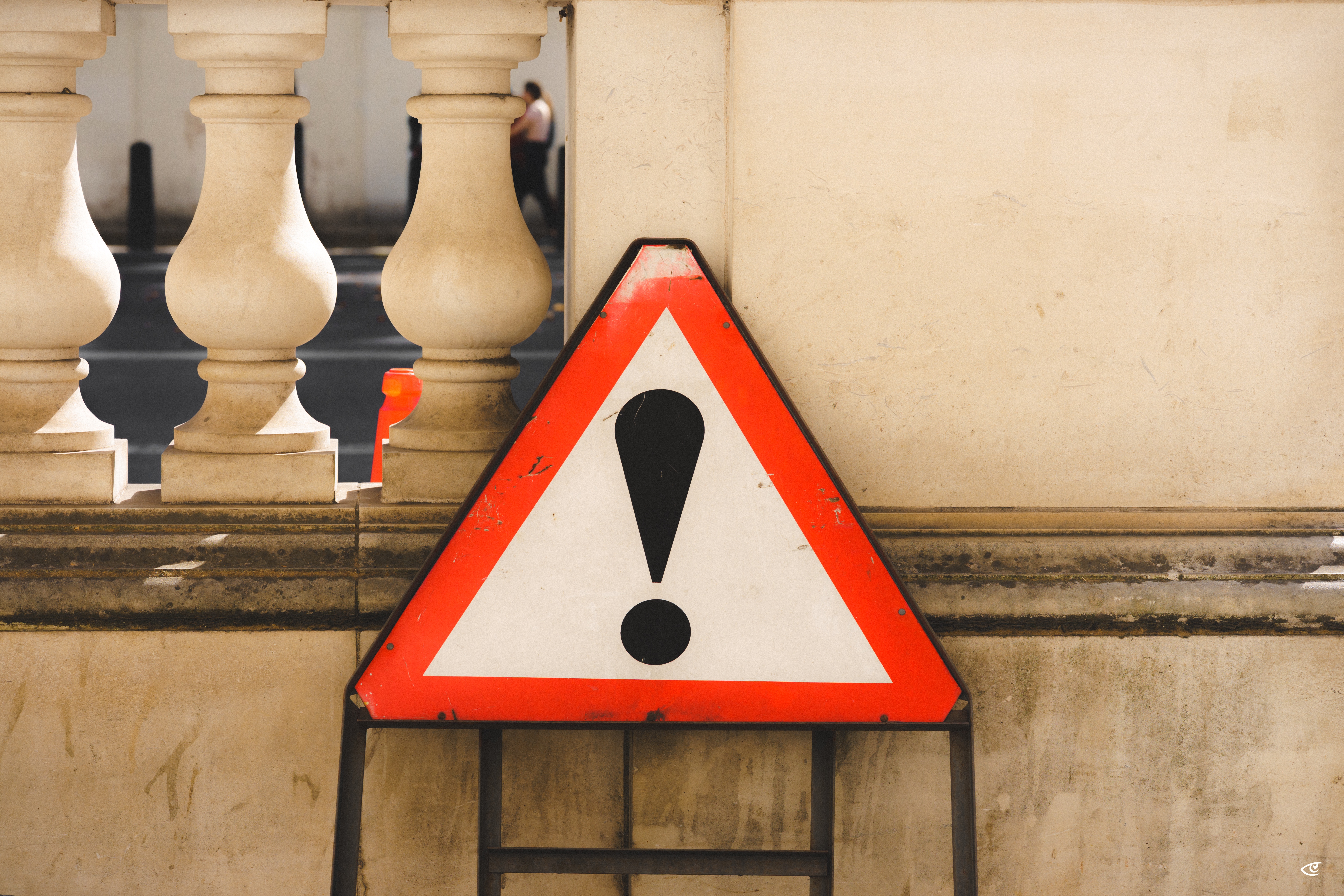 Red-and-white triangular warning sign with a black exclamation mark leaning against a beige stone wall beside a balustrade, with street and a blurred pedestrian in the background.