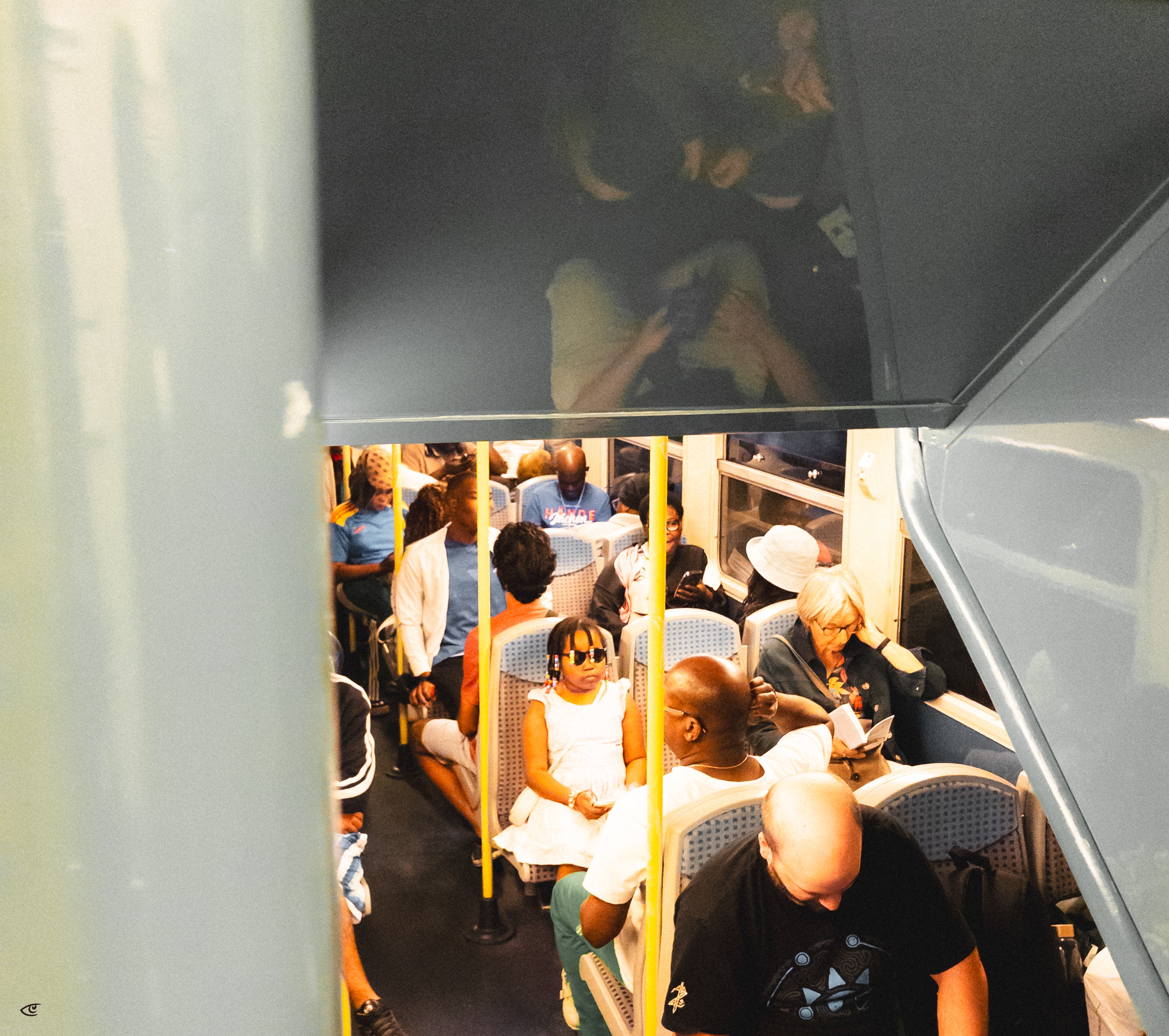 View down the aisle of a crowded subway car with blue seats and yellow poles; passengers sit and stand, including a child in a white dress and sunglasses near the center, while others look at phones or read by the windows.