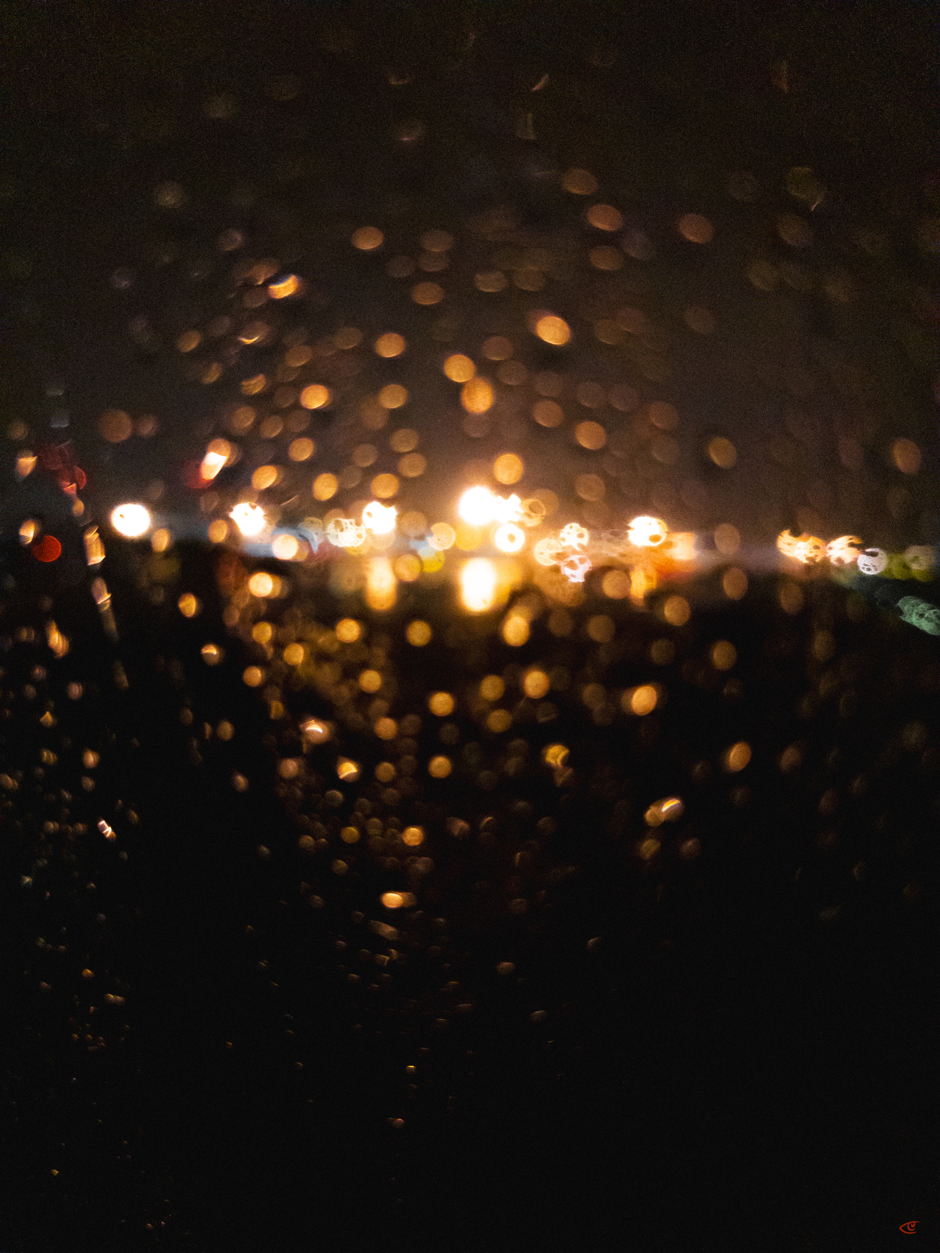 Out-of-focus nighttime streetlights seen through a rain-speckled window, with glowing bokeh circles across the center and dark diagonal bands in the foreground.