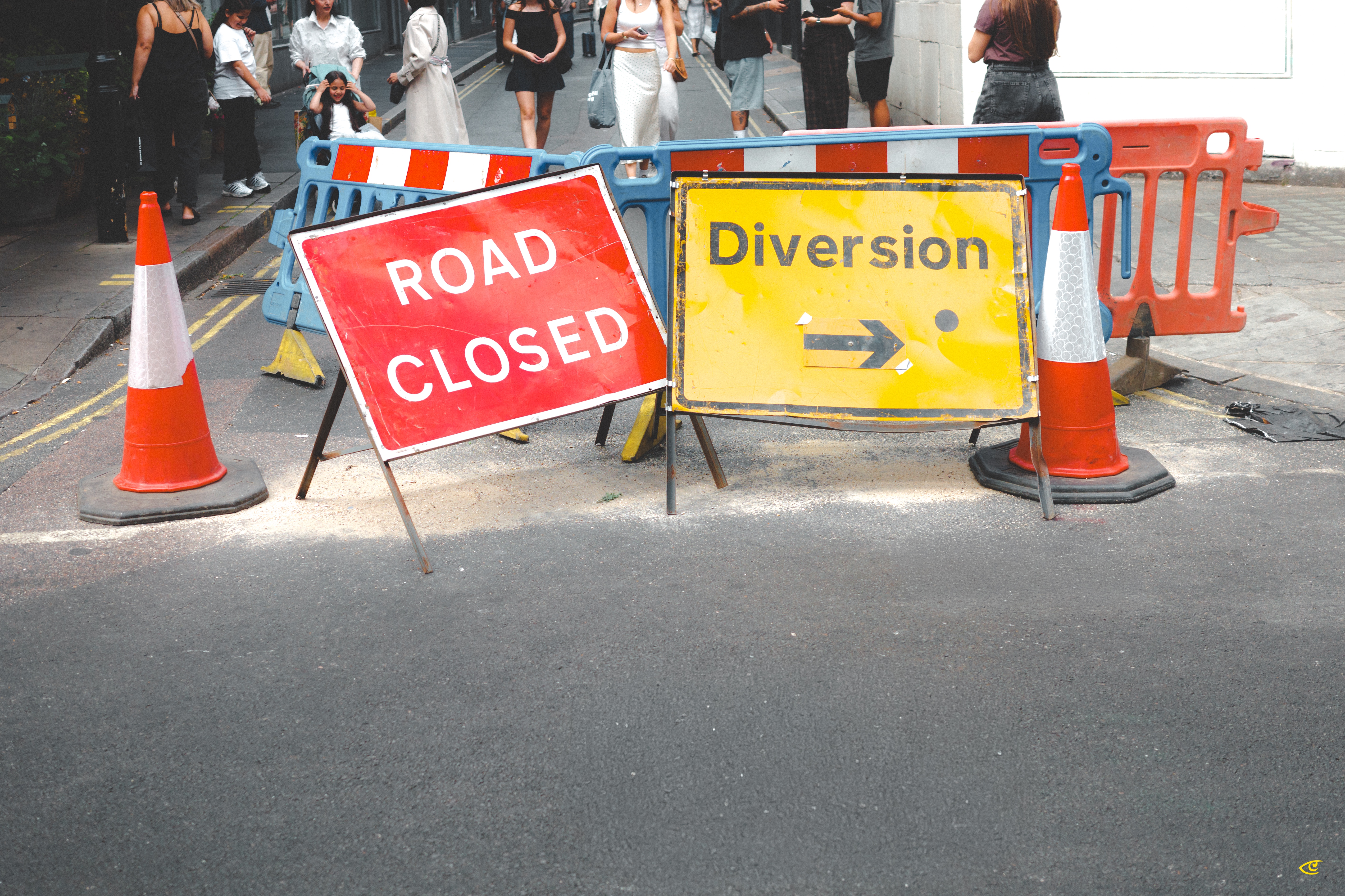 Red “ROAD CLOSED” sign and yellow “Diversion” sign with a right-pointing arrow placed in front of blue and orange plastic barriers, flanked by orange traffic cones on a city street with pedestrians in the background.