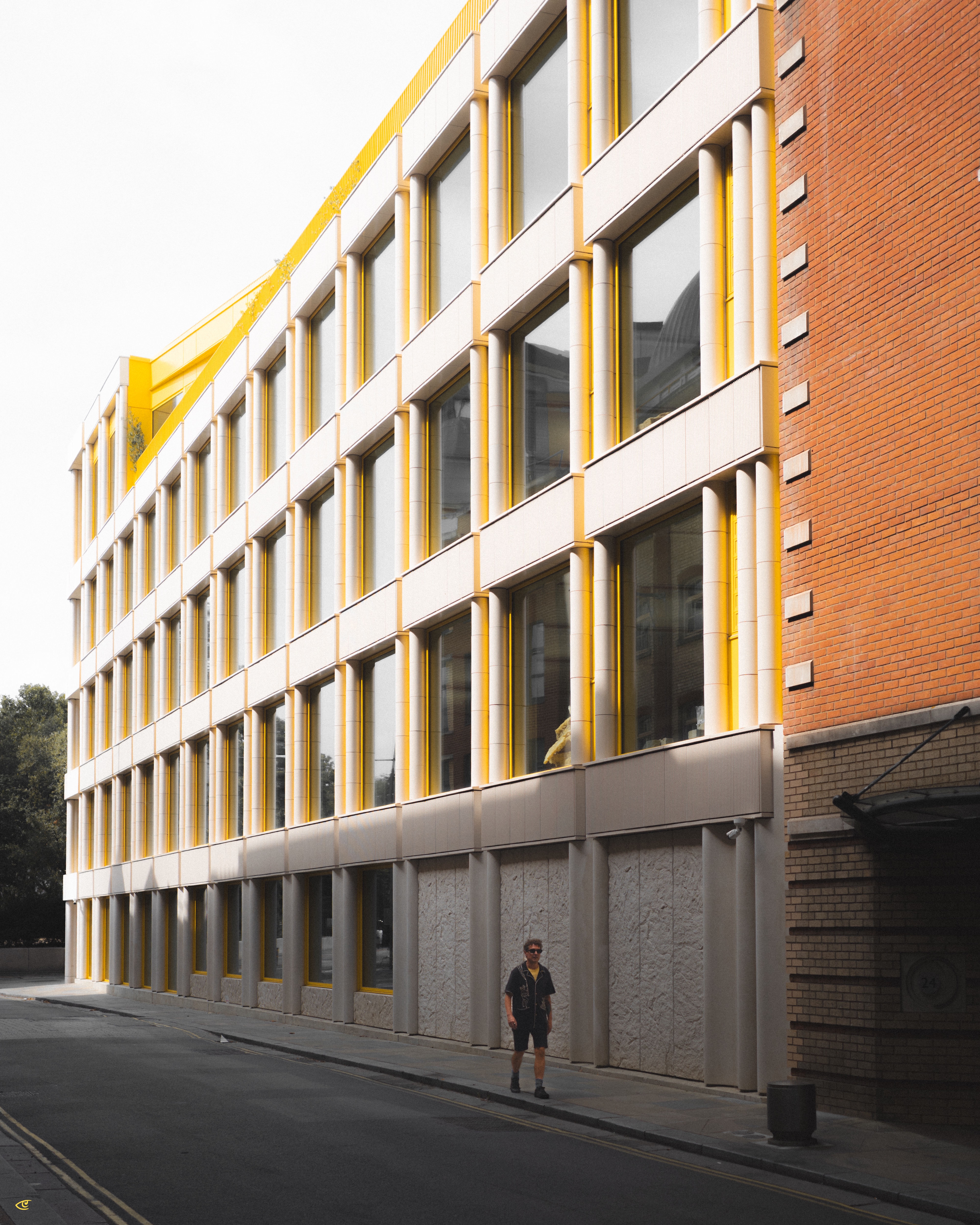 Angled view down a quiet street beside a multi-story modern building with large rectangular windows framed by yellow vertical columns and light gray panels; a single pedestrian walks on the sidewalk near the base, with a brick wall section on the right edge.