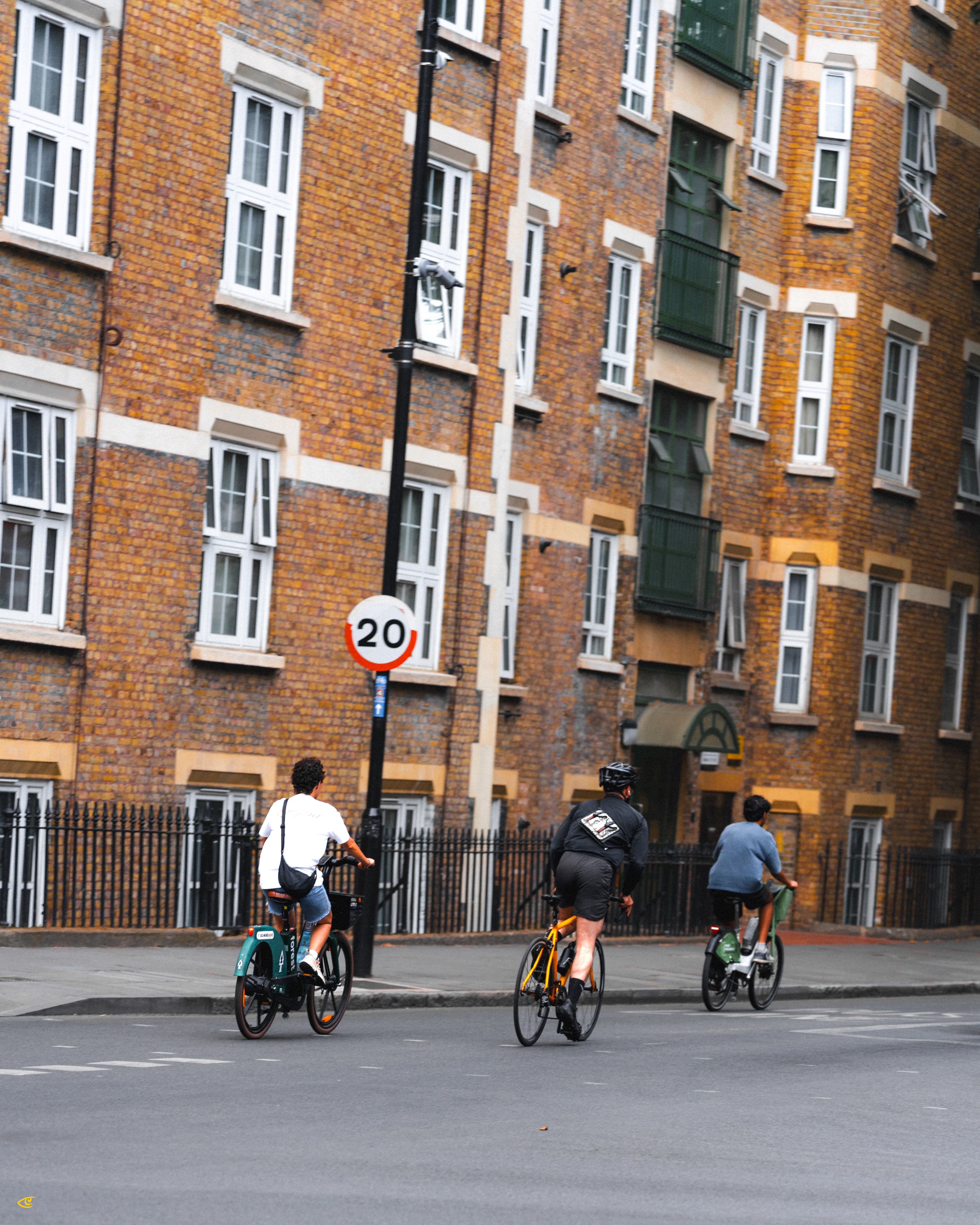 Three cyclists riding along a city street in front of a multi-story brick apartment building with many white-framed windows; a pole with a circular “20” speed limit sign stands near the curb.