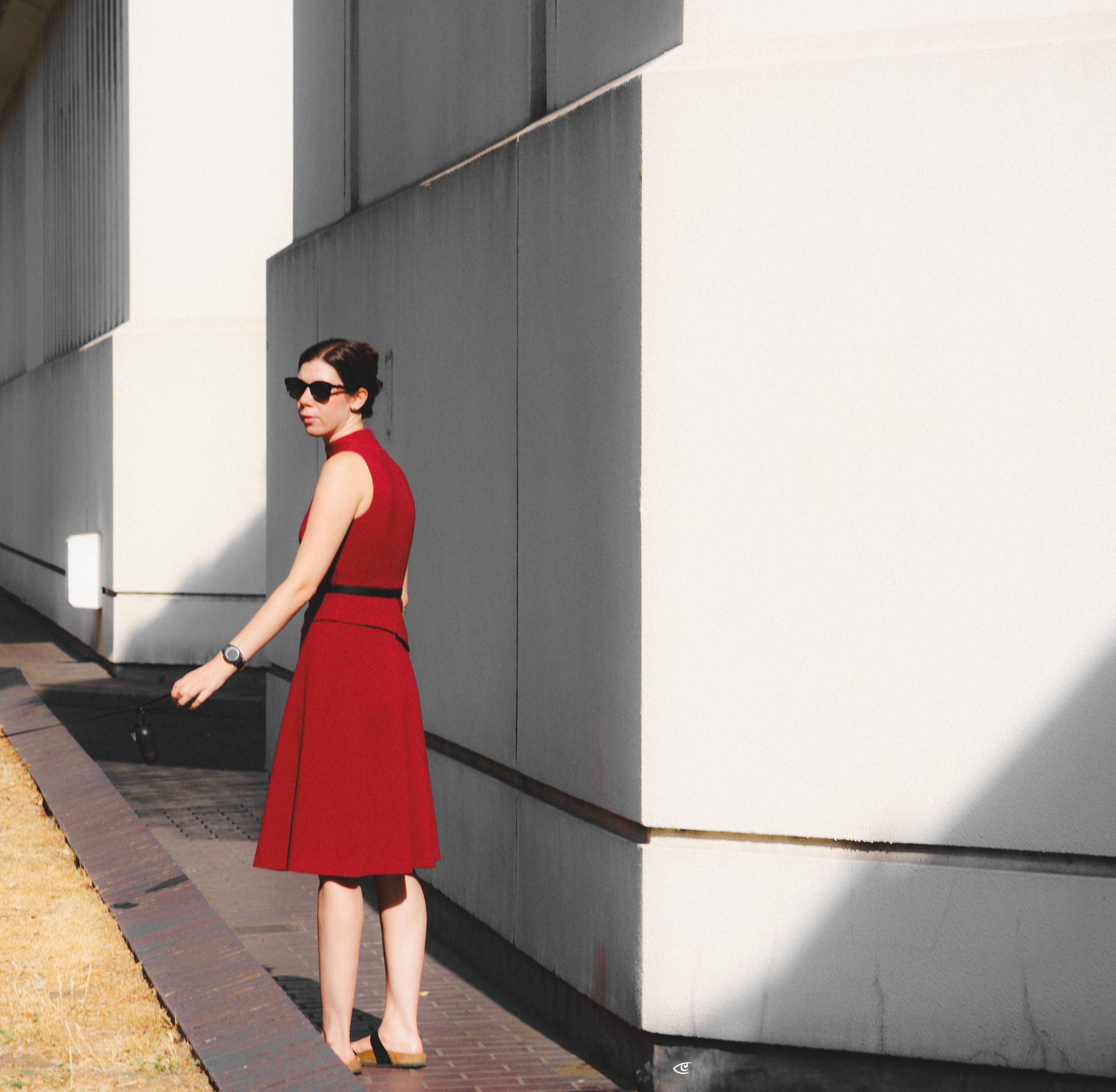 Woman in a sleeveless red dress and sunglasses standing on a narrow sidewalk beside a modern white-and-gray building, with long diagonal shadows across the wall and ground.