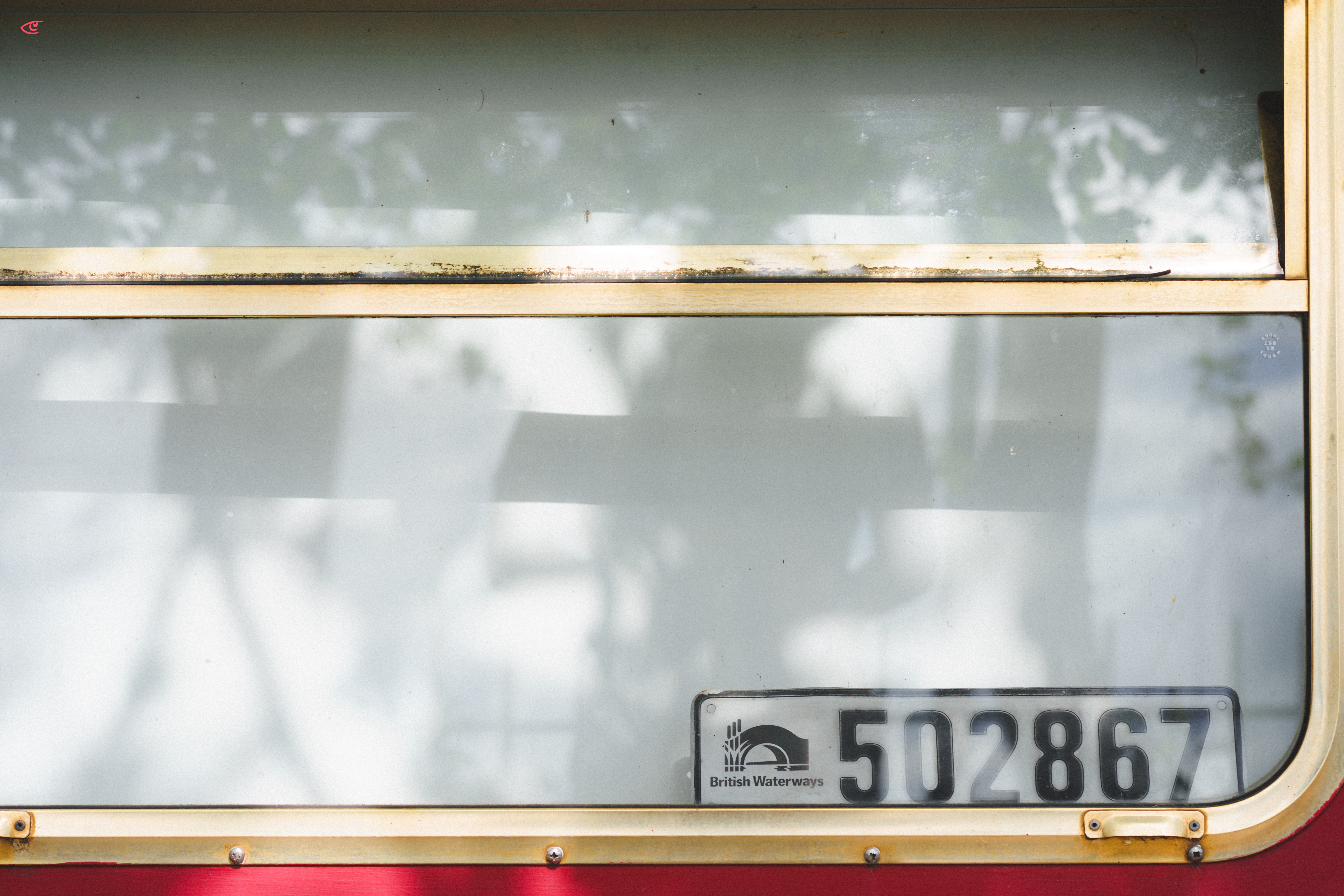 Close-up of a vehicle window with metal trim; a “British Waterways” license plate reading “502867” is visible behind the glass at the lower right, with faint tree reflections across the window and a red panel along the bottom edge.
