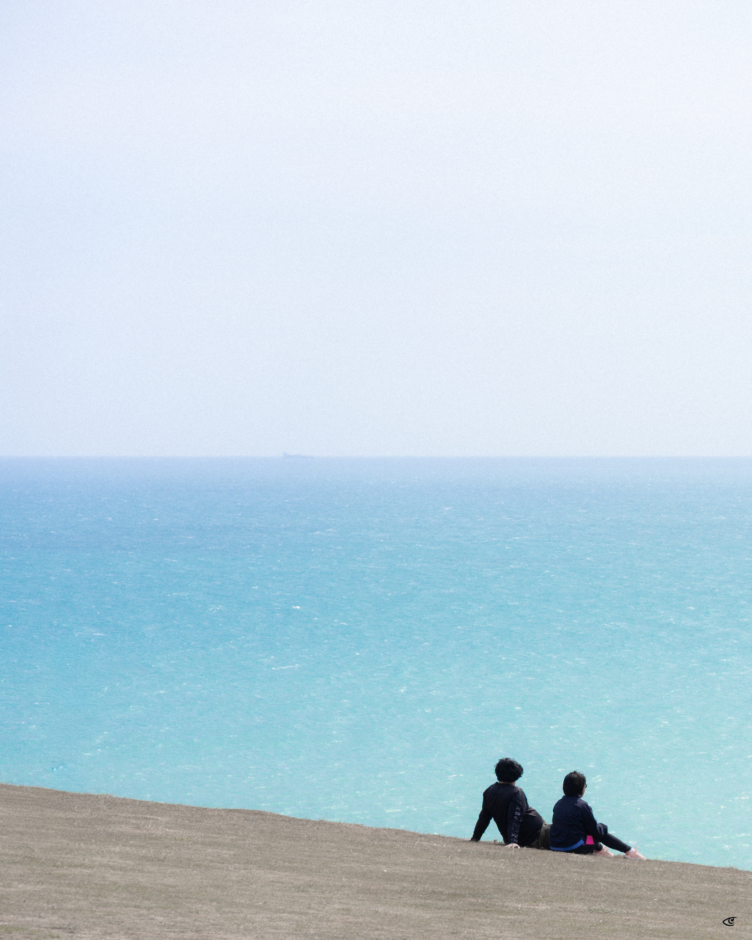 Two people sit on a sandy slope in the lower right, facing a turquoise ocean under a pale sky with the horizon across the middle of the frame.