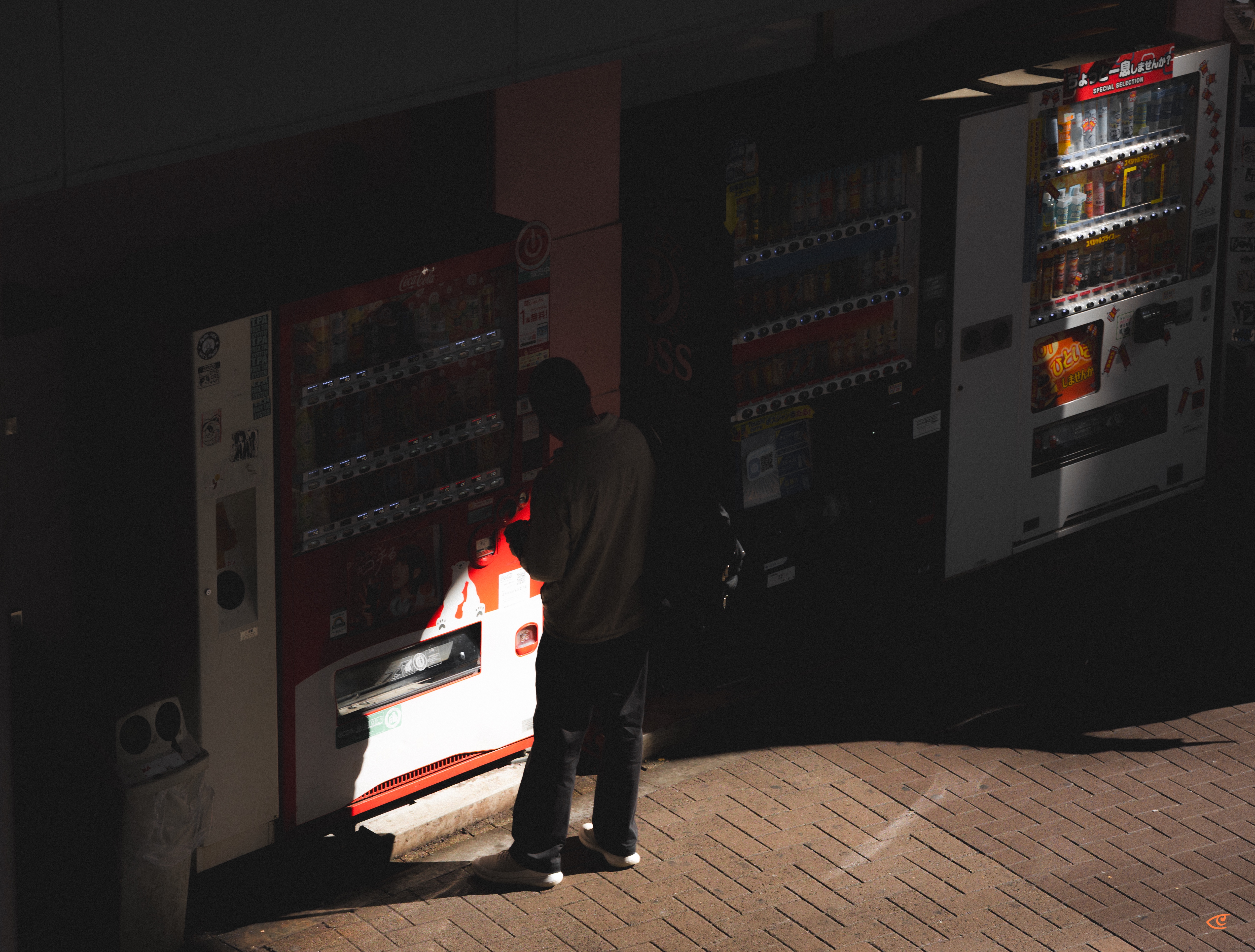Person standing on a brick-paved sidewalk using a red vending machine in a dimly lit row of vending machines, with the machine’s bright light illuminating the person and casting a shadow; additional machines line the wall to the right.