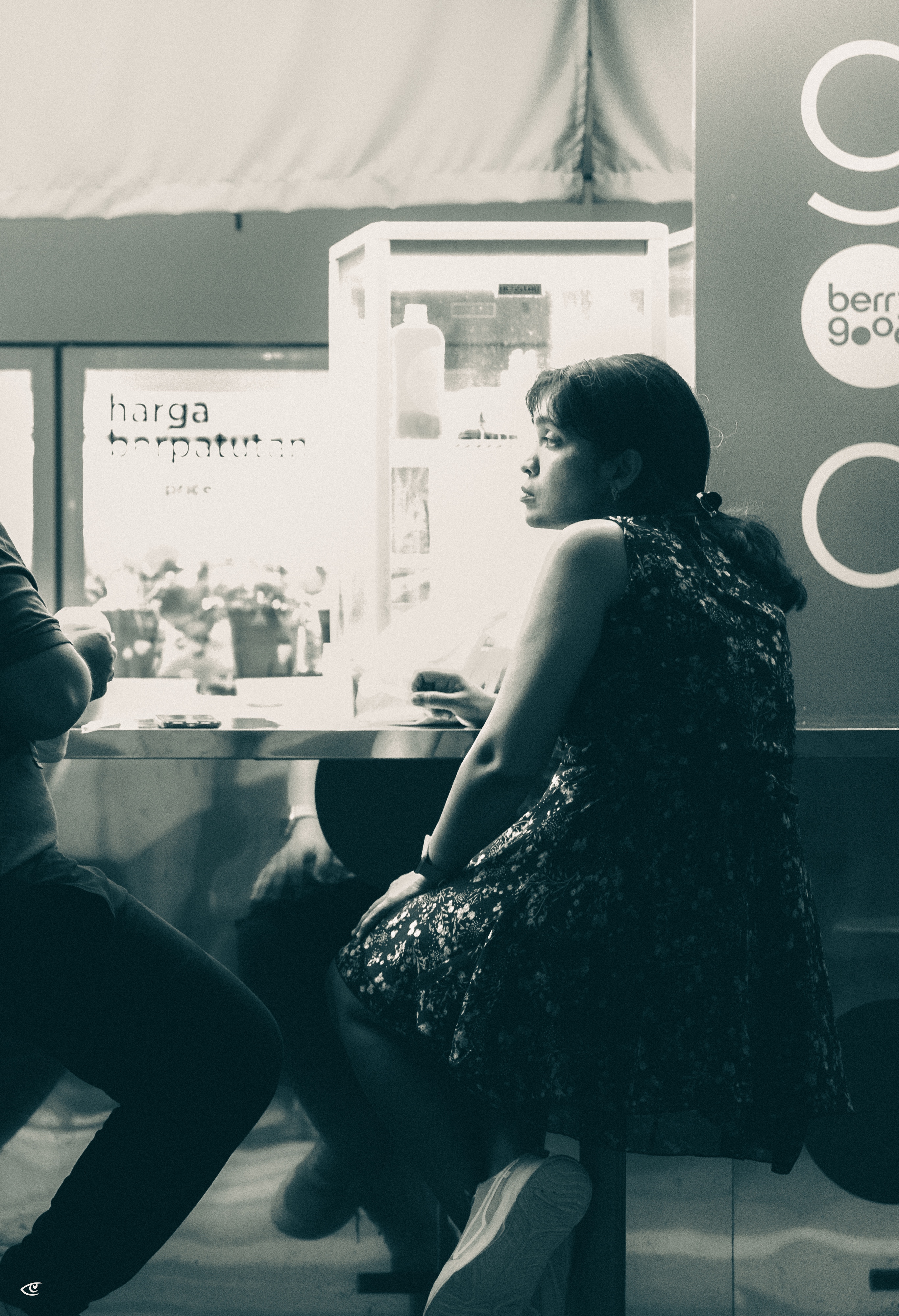 Woman in a patterned sleeveless dress sits sideways on a stool at a café counter, holding a drink and looking to the right; another person is partially visible on the left, with a small refrigerator and signage behind them.