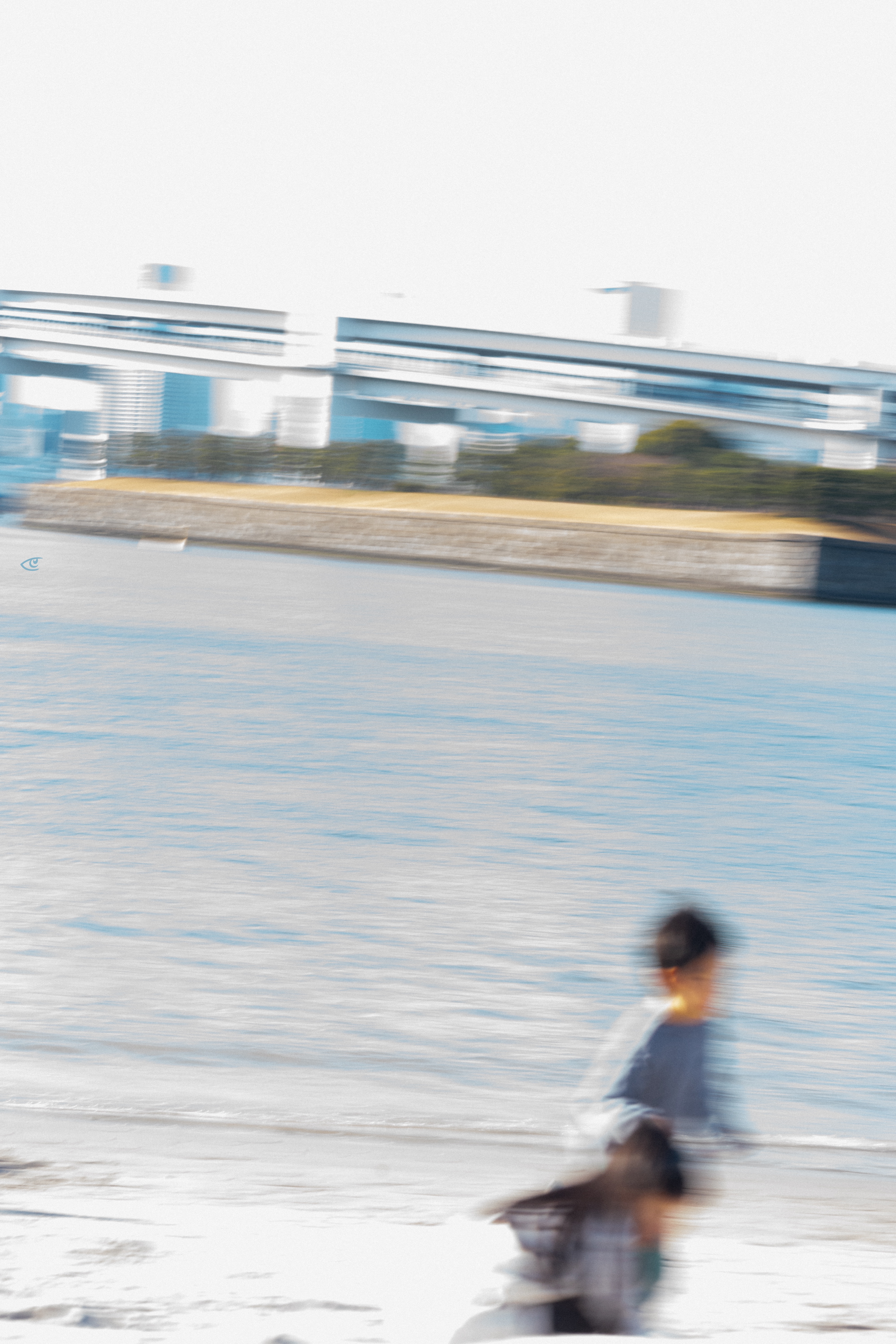 Blurred view of a waterfront with rippling water and a sandy shoreline in the foreground; two motion-blurred people near the bottom right, and an elevated bridge with distant buildings across the water in the background.