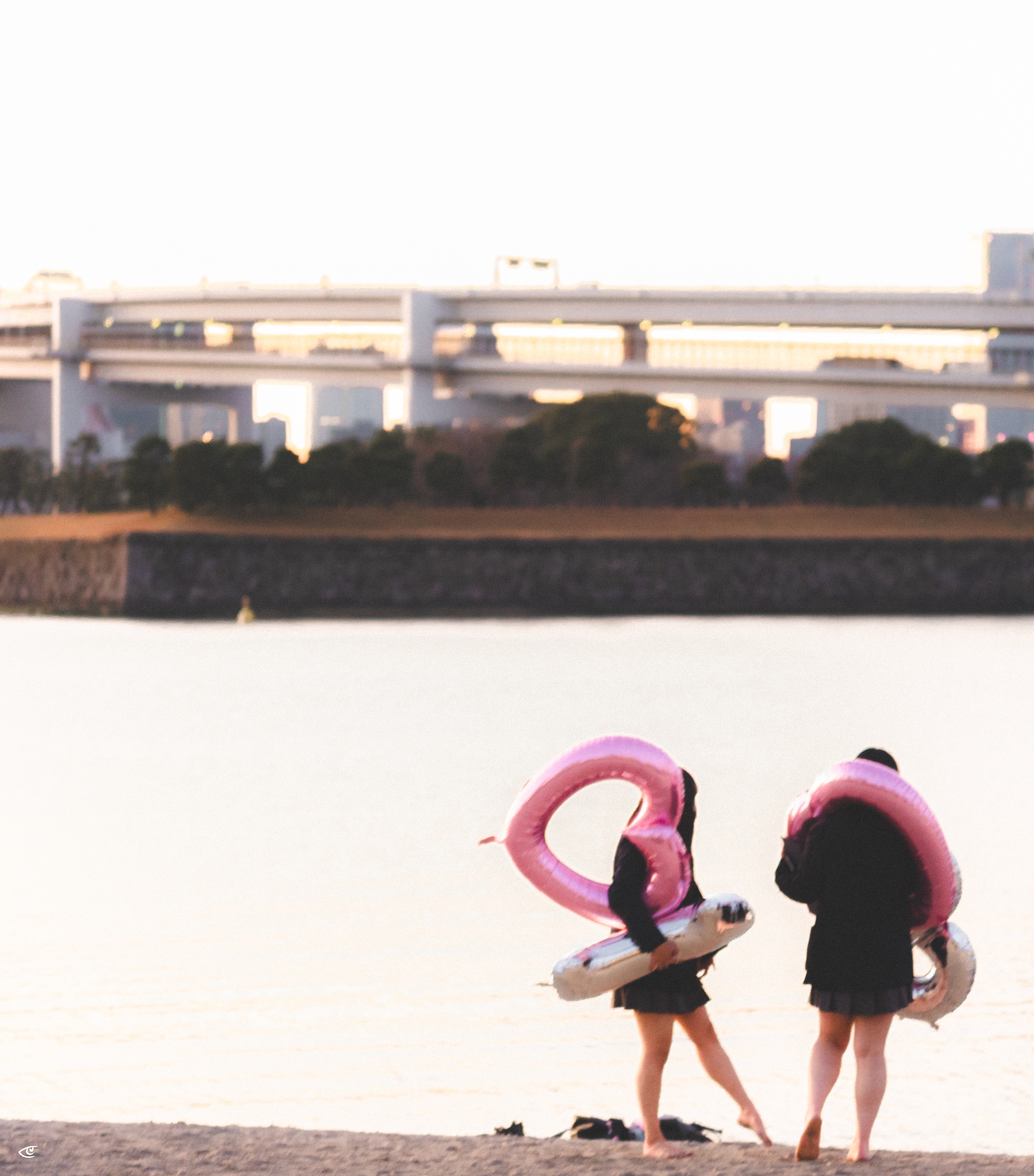 Two people standing on a sandy shore facing the water, each carrying a large pink-and-white inflatable swim ring; a distant bridge and city buildings are visible across the water.