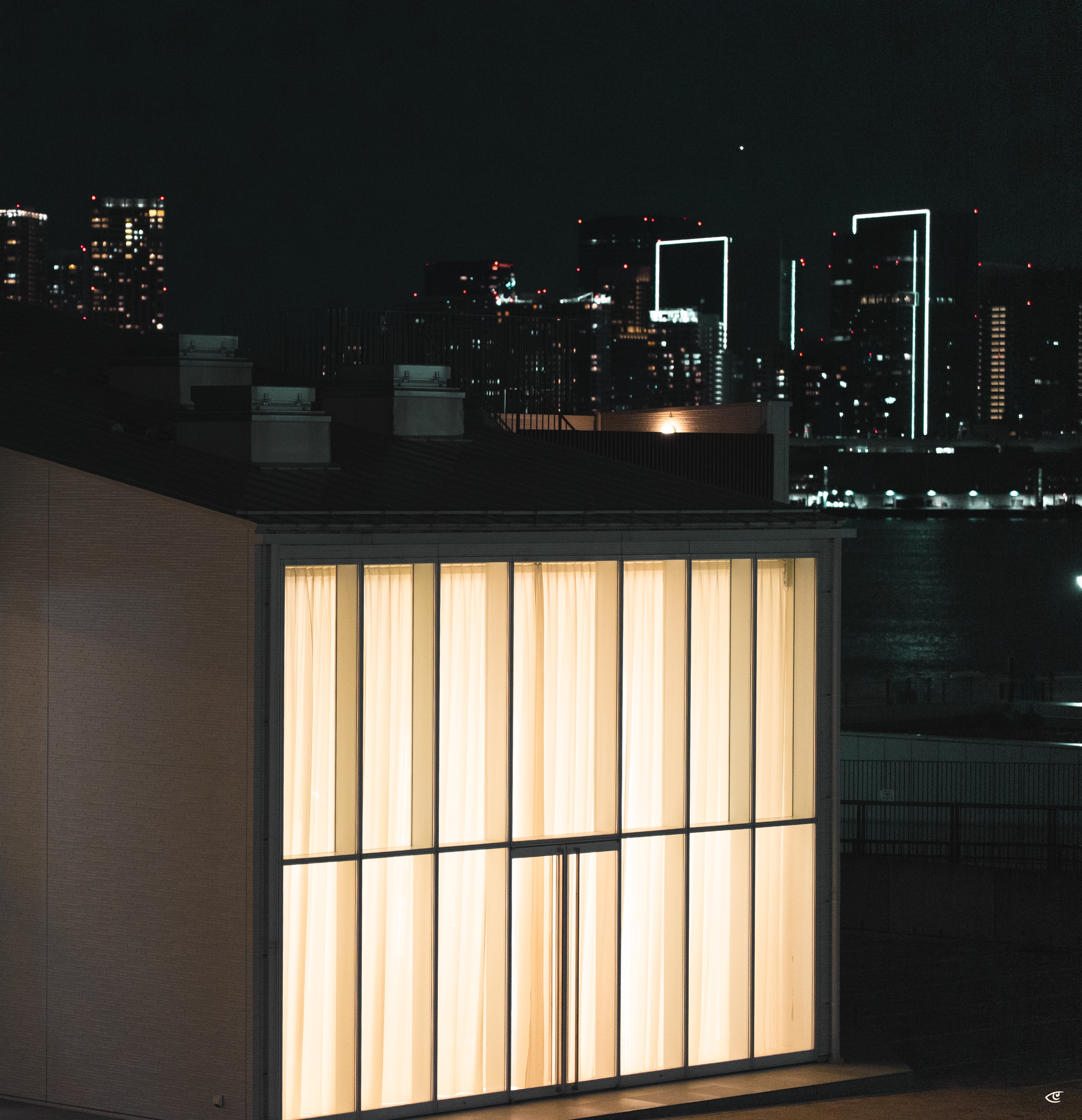 Large illuminated window with sheer curtains on a low building in the foreground, with a dark river and distant city skyline at night behind it, including high-rises outlined in white lights.