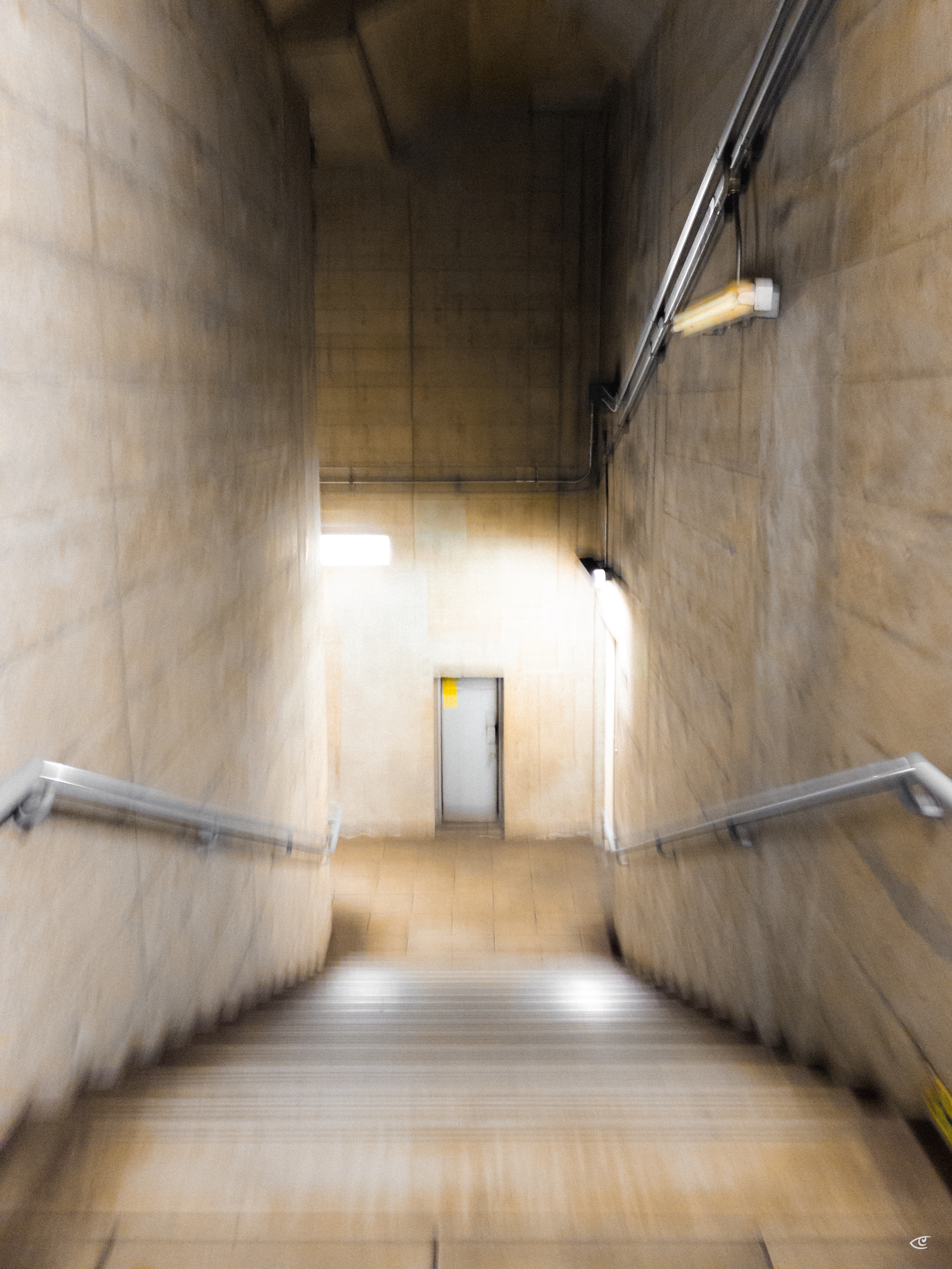 Concrete stairwell with handrails on both sides leading down to a closed gray door; overhead pipes and fluorescent lights line the right wall, and the image is slightly motion-blurred.