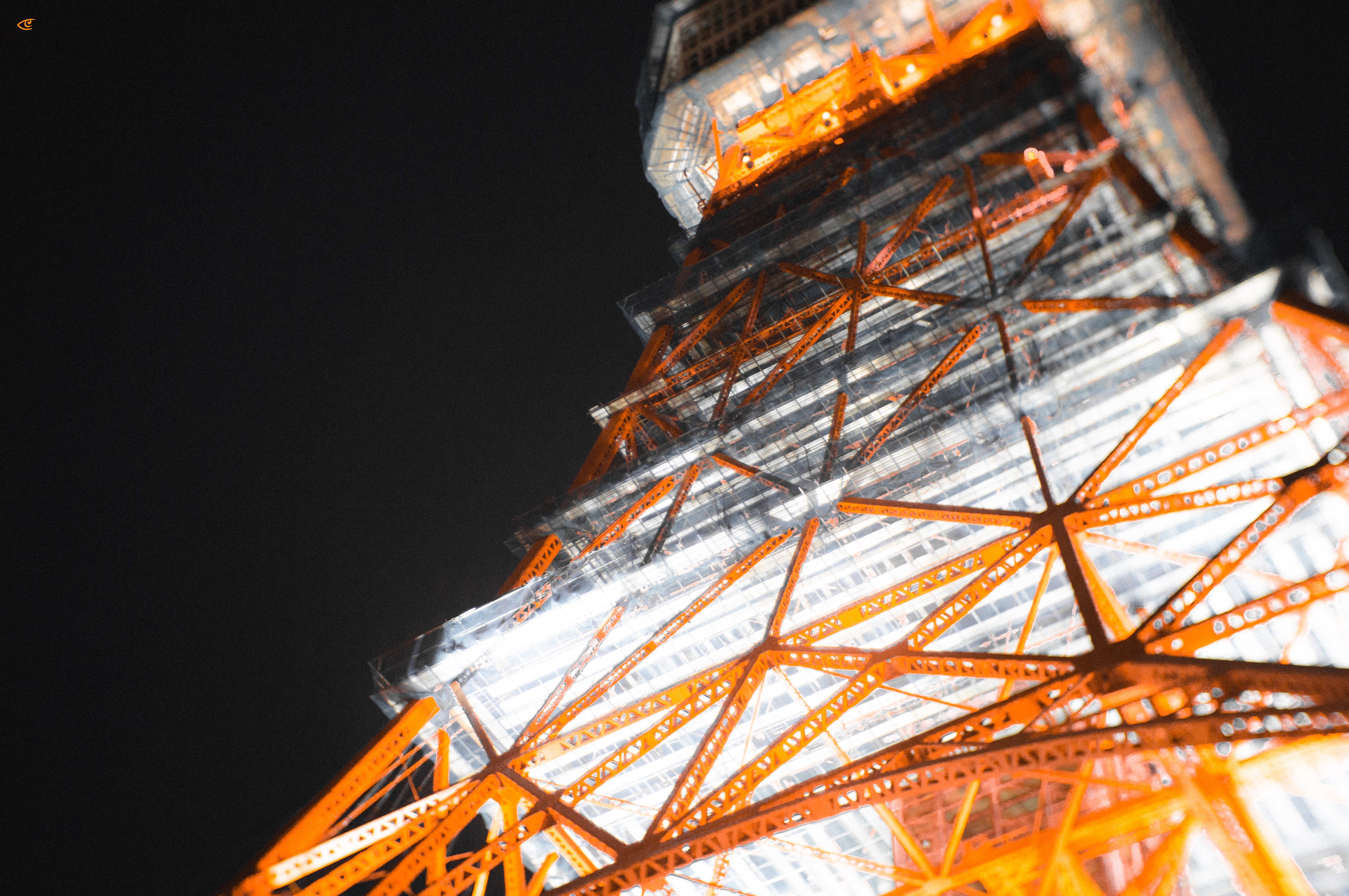Upward view of an orange-and-white steel lattice tower illuminated at night, with bright lights along the structure against a dark sky.