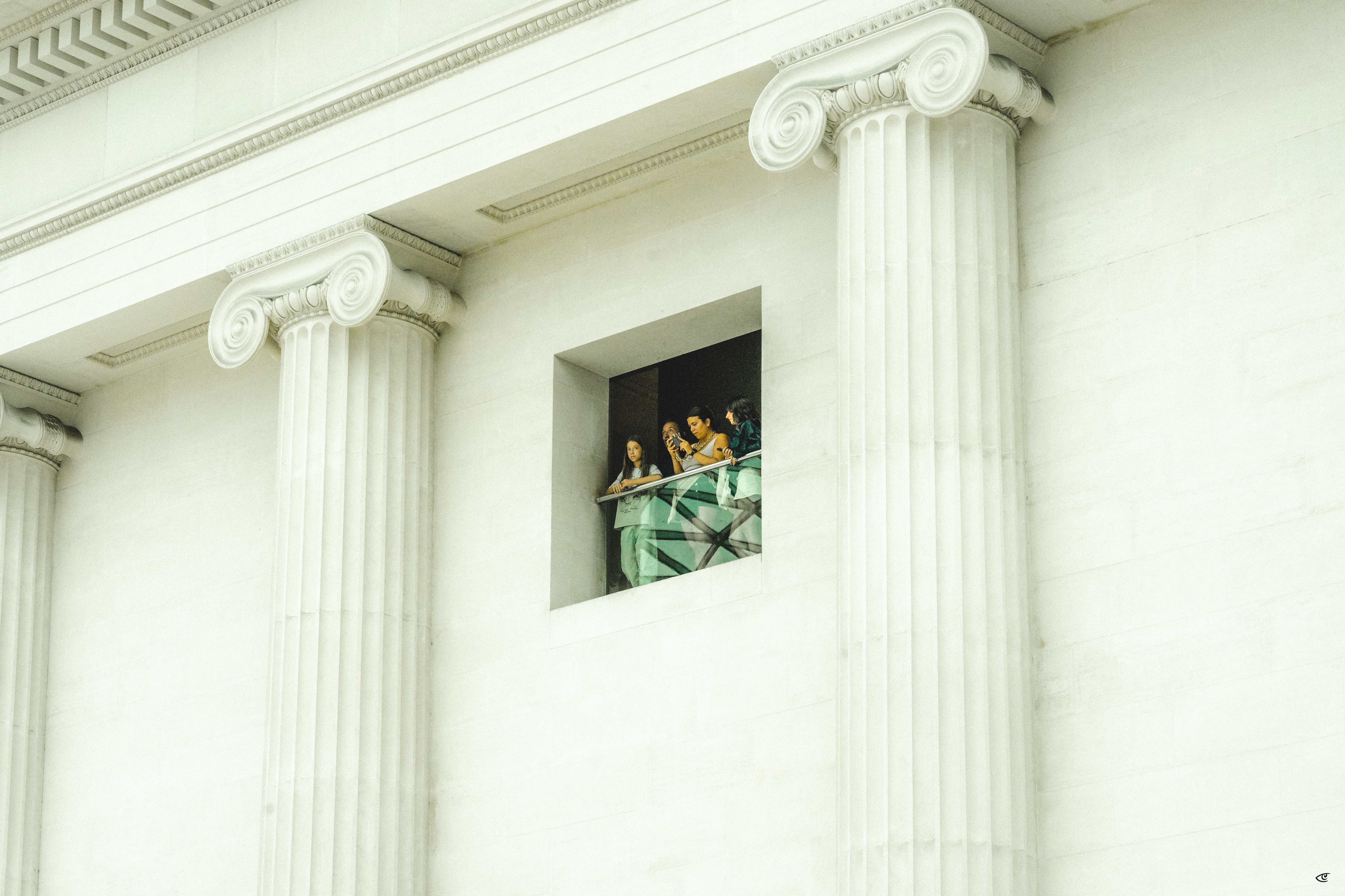 Group of people standing on a small balcony inside a square window opening between two large white fluted columns on a neoclassical building facade.