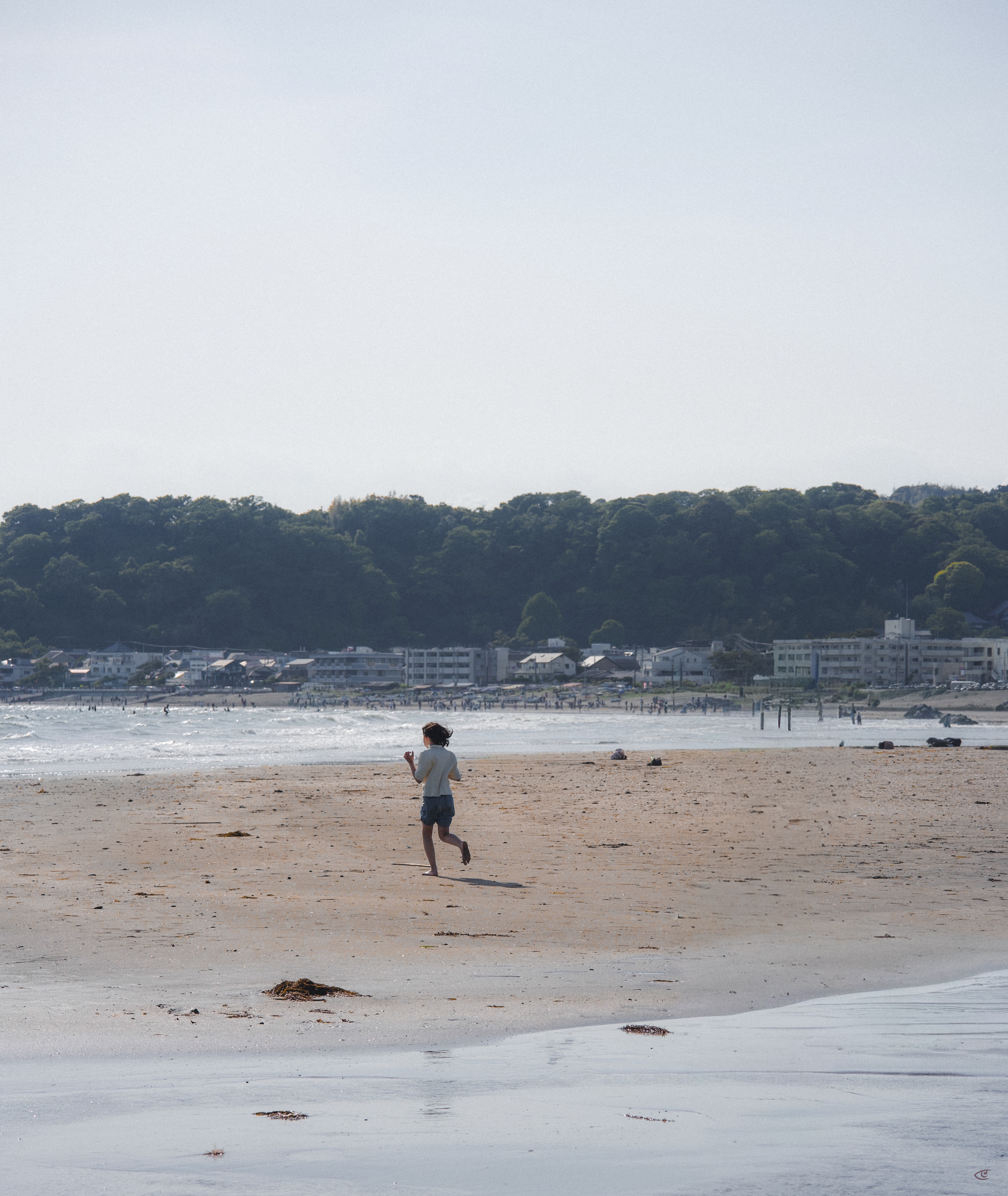 Person running barefoot on a wide sandy beach near the shoreline, with gentle waves, scattered seaweed, distant beachgoers, low buildings along the coast, and a forested hillside under a pale sky.