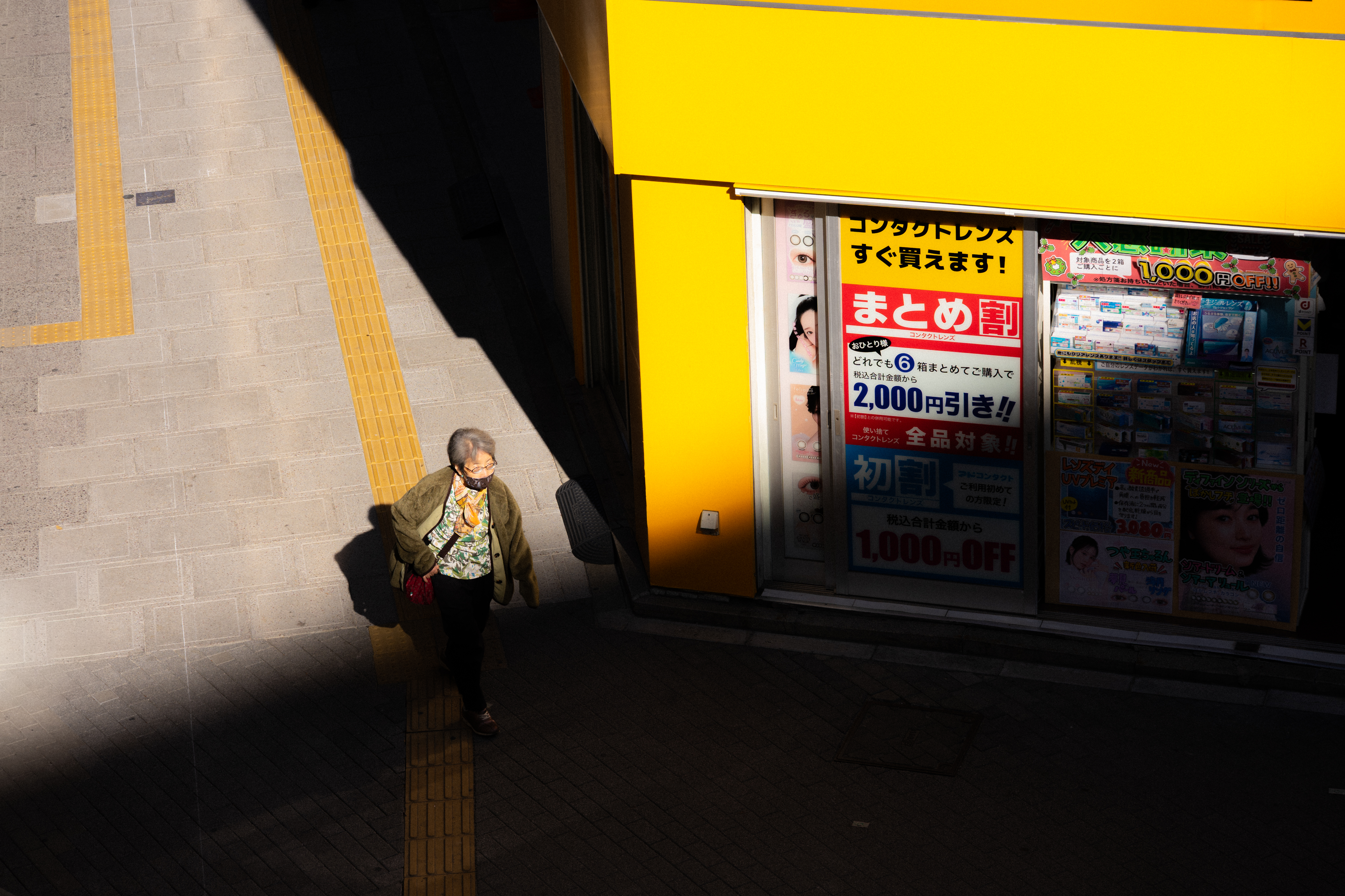 Person walking on a shadowed street next to a brightly-lit building with a yellow exterior. The building features a large window displaying colorful posters with Japanese text. A tiled walkway with a yellow tactile guidance path is visible.