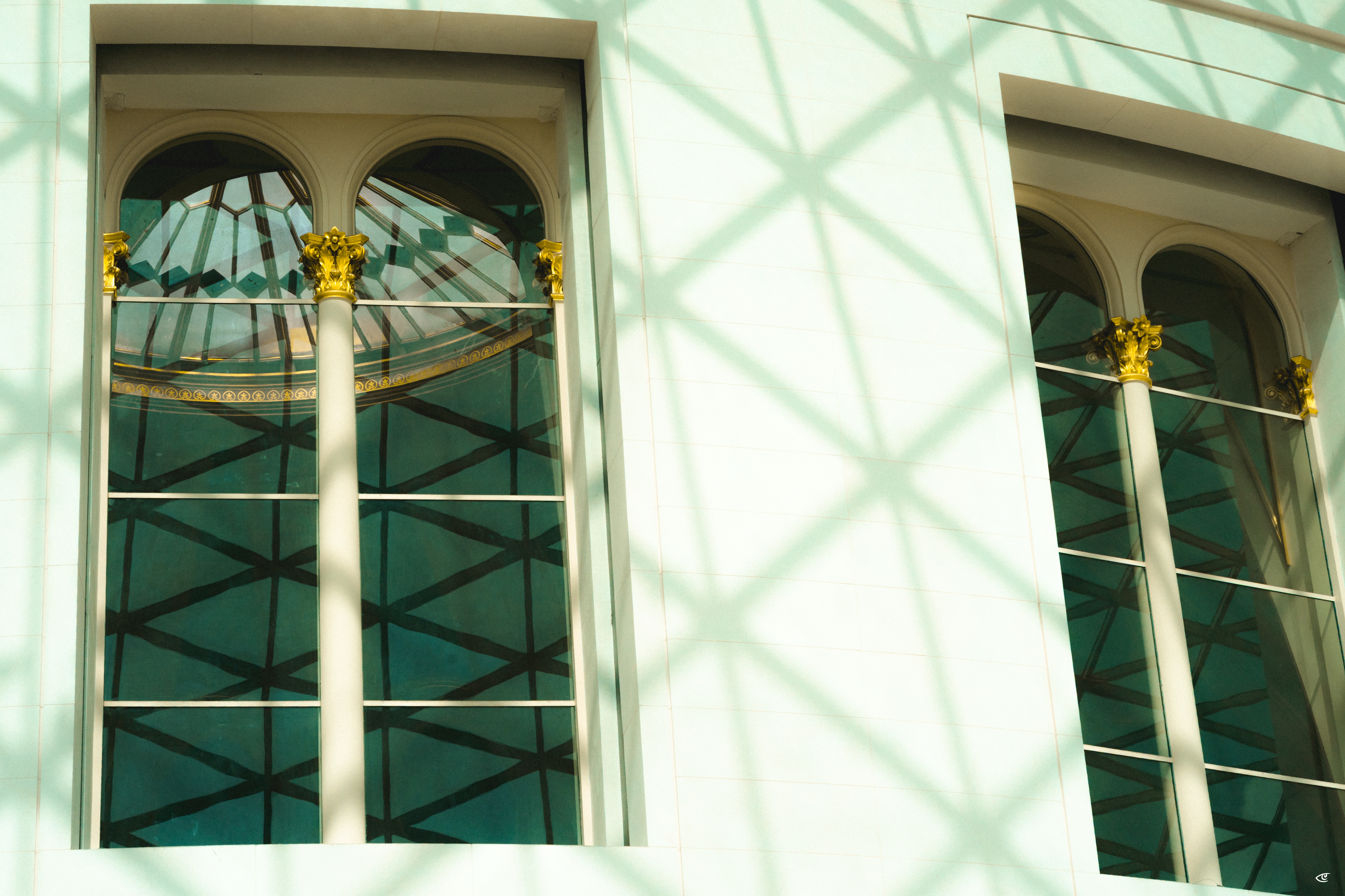 Two tall arched windows on a light-colored building facade, framed by columns with gold capitals; teal glass reflects a geometric dome pattern, with diagonal lattice shadows cast across the wall.