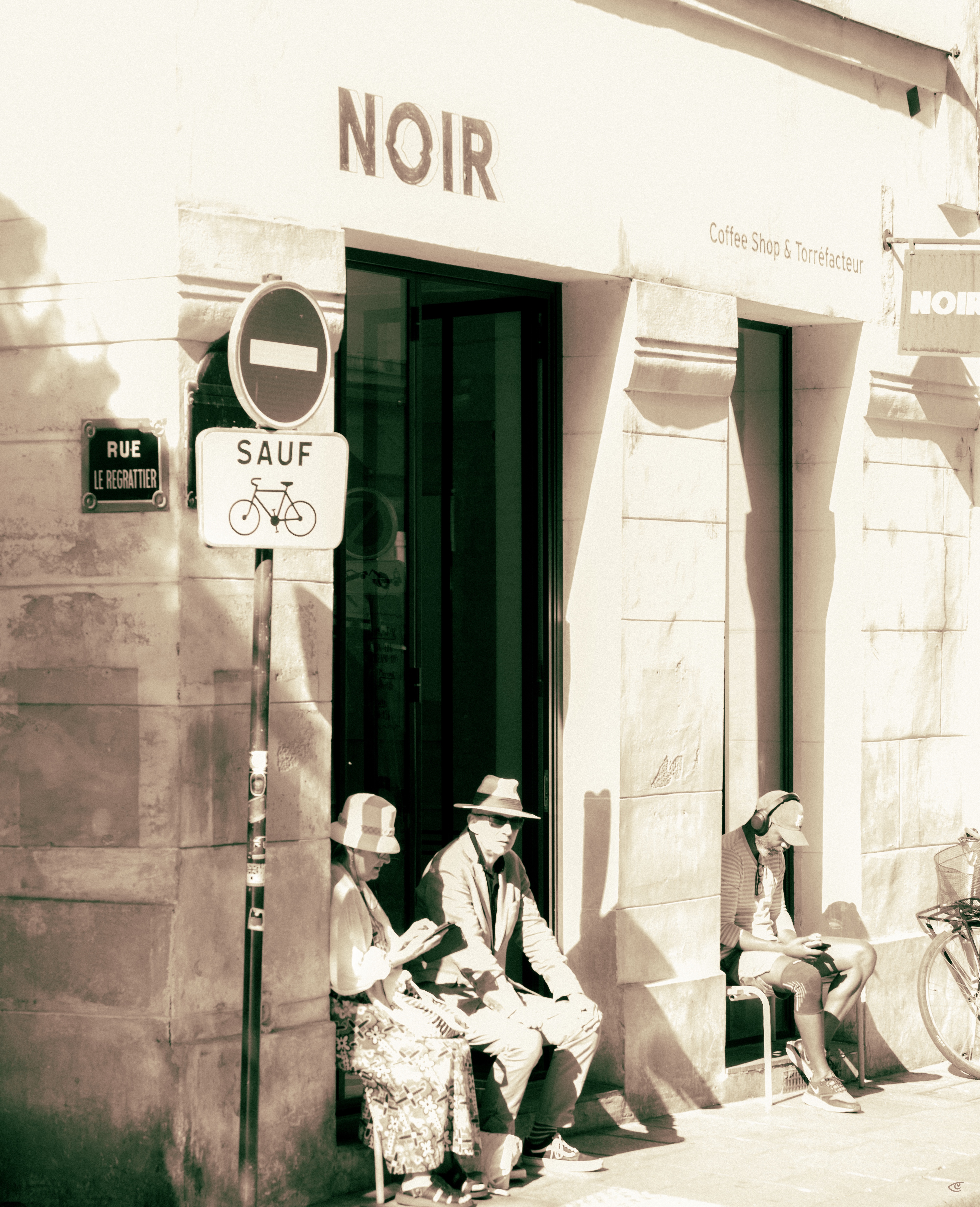 Street corner café storefront with “NOIR” sign above a doorway and “Coffee Shop & Torréfacteur” on the wall; a no-entry sign with “SAUF” and a bicycle icon stands beside a “RUE LE REGRATTIER” plaque. Three people sit along the wall in sunlight—two near the doorway and one in a recessed entry—while a bicycle wheel is partially visible at the right edge.