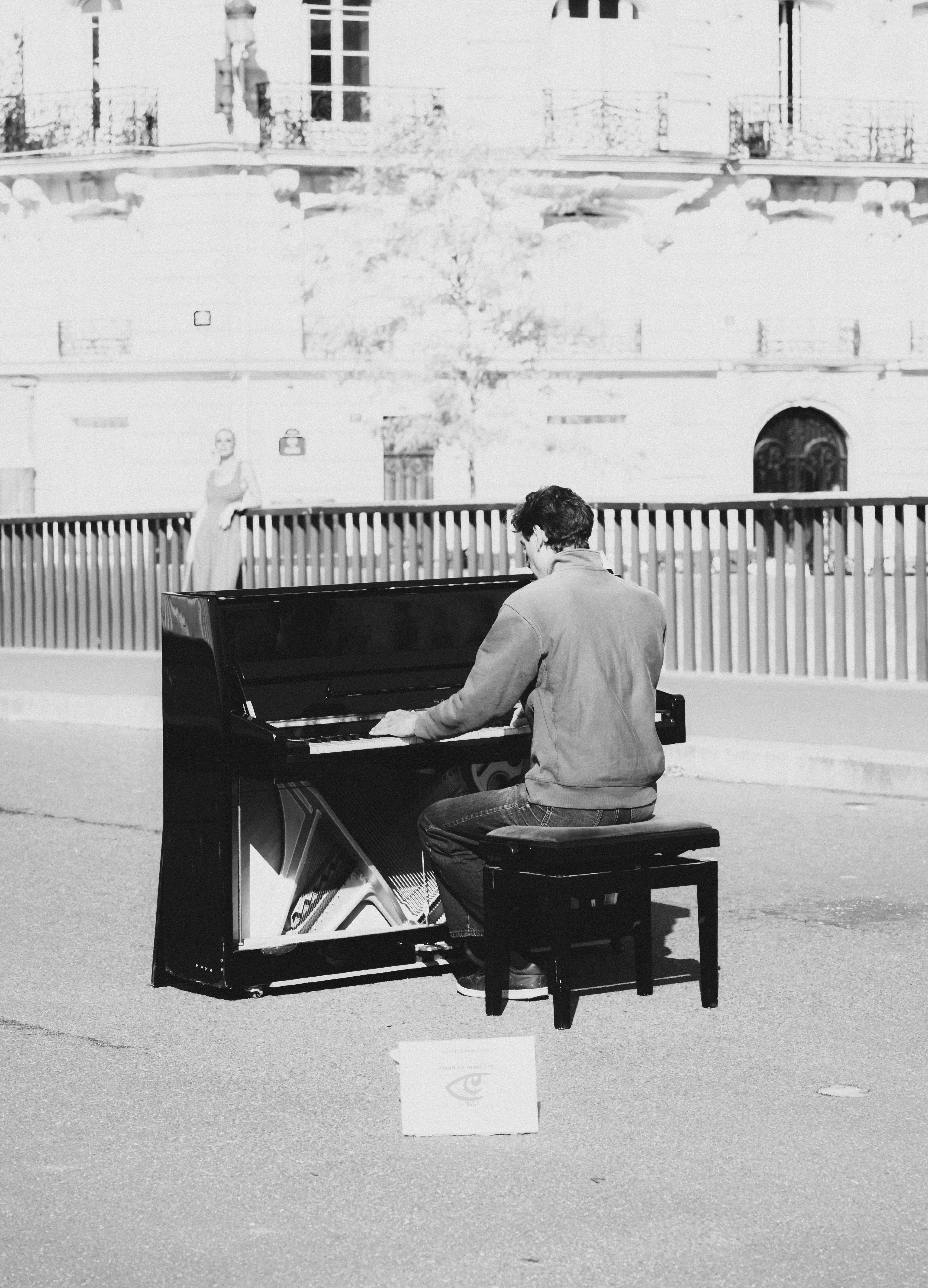 Black-and-white photo of a man seated on a bench playing an upright piano placed on a paved street, with a small sign on the ground in front; a railing and a large building facade with windows in the background, and a person standing behind the railing.