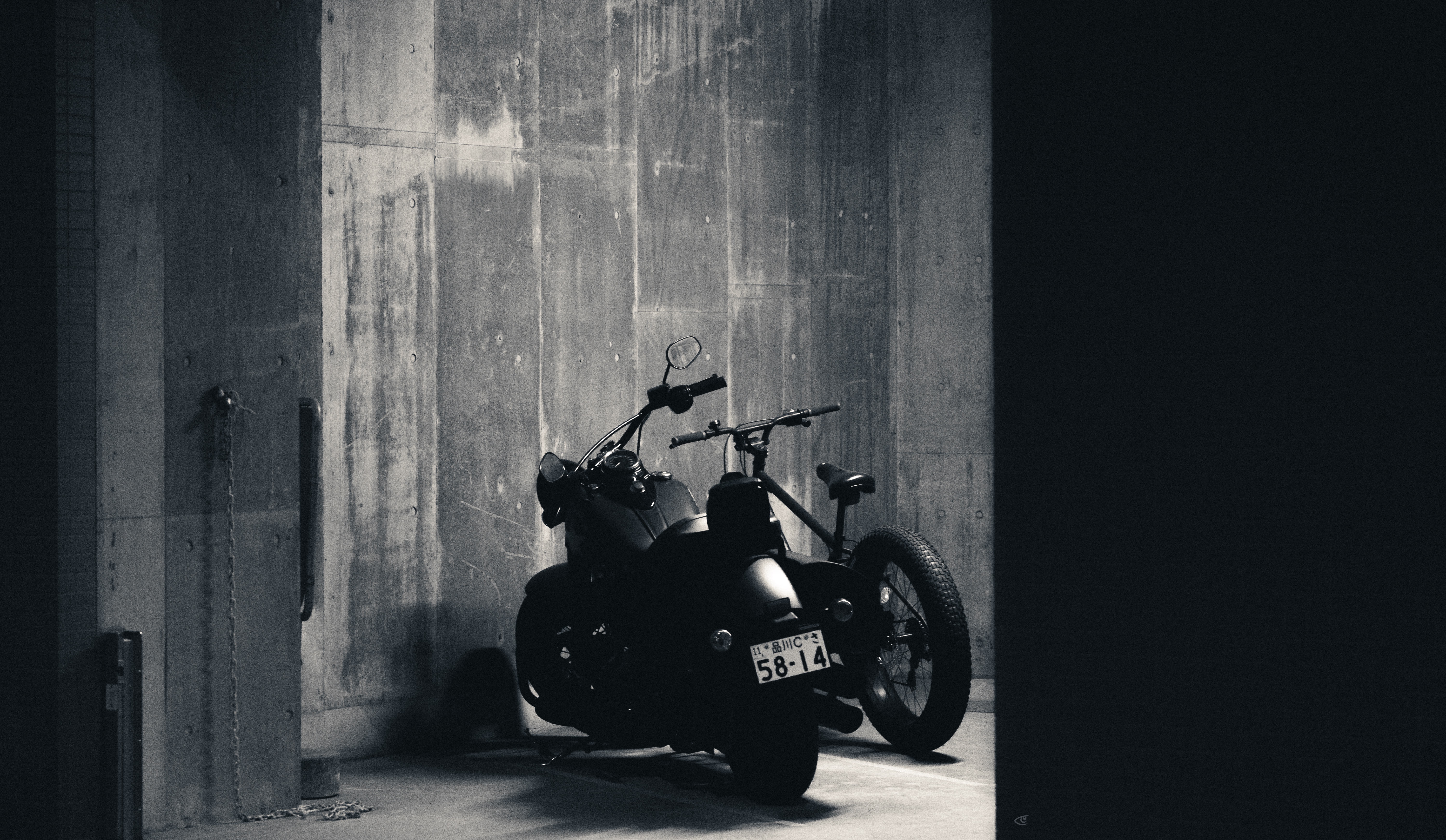 Two motorcycles parked side by side in a dim concrete garage, viewed from a doorway with strong shadows and a partly visible license plate.
