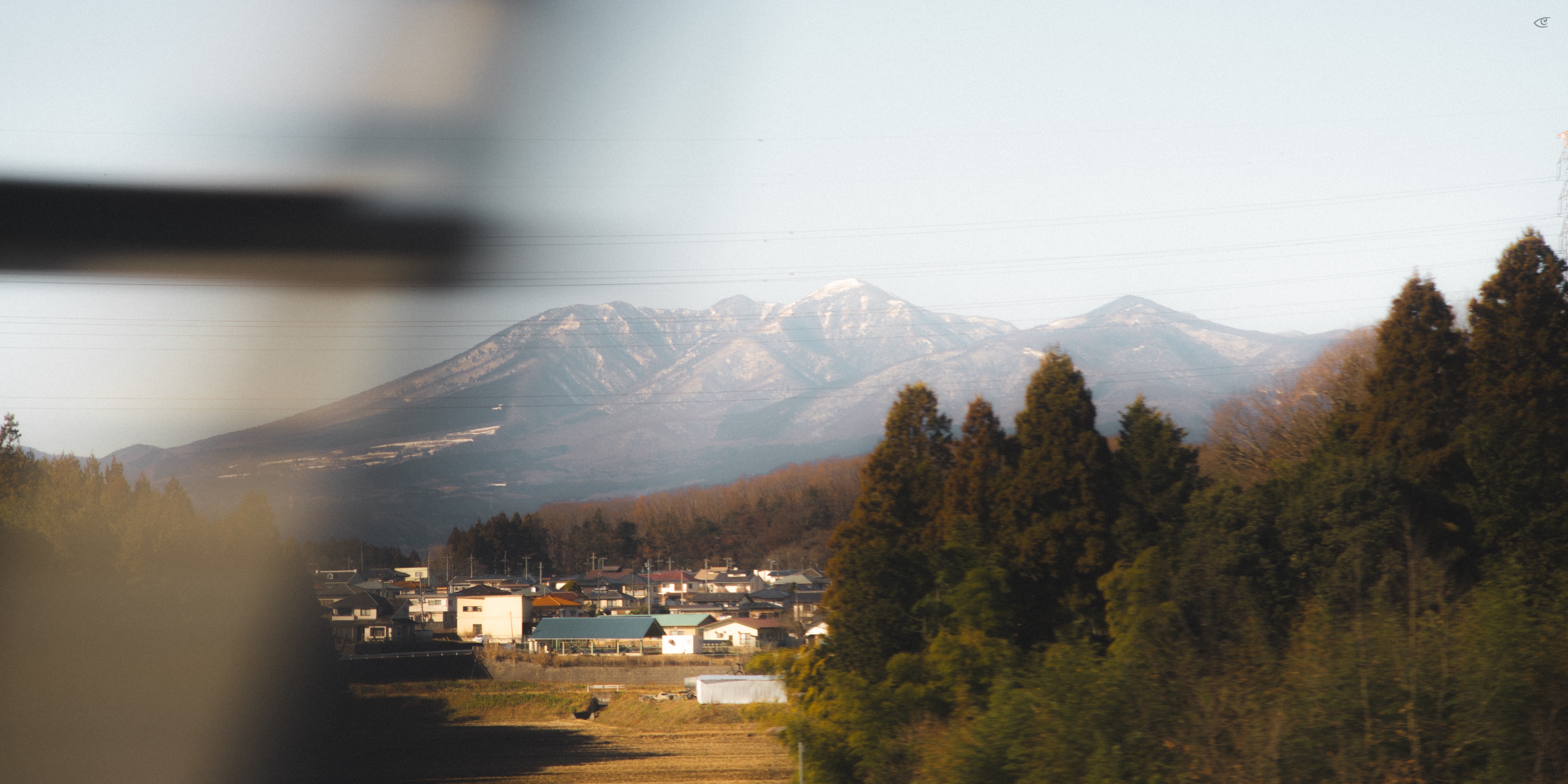 Snow-dusted mountain range in the background, with a small town and fields in the valley below and a cluster of tall evergreen trees on the right; blurred dark shapes and a horizontal streak frame the left side and upper portion of the image.