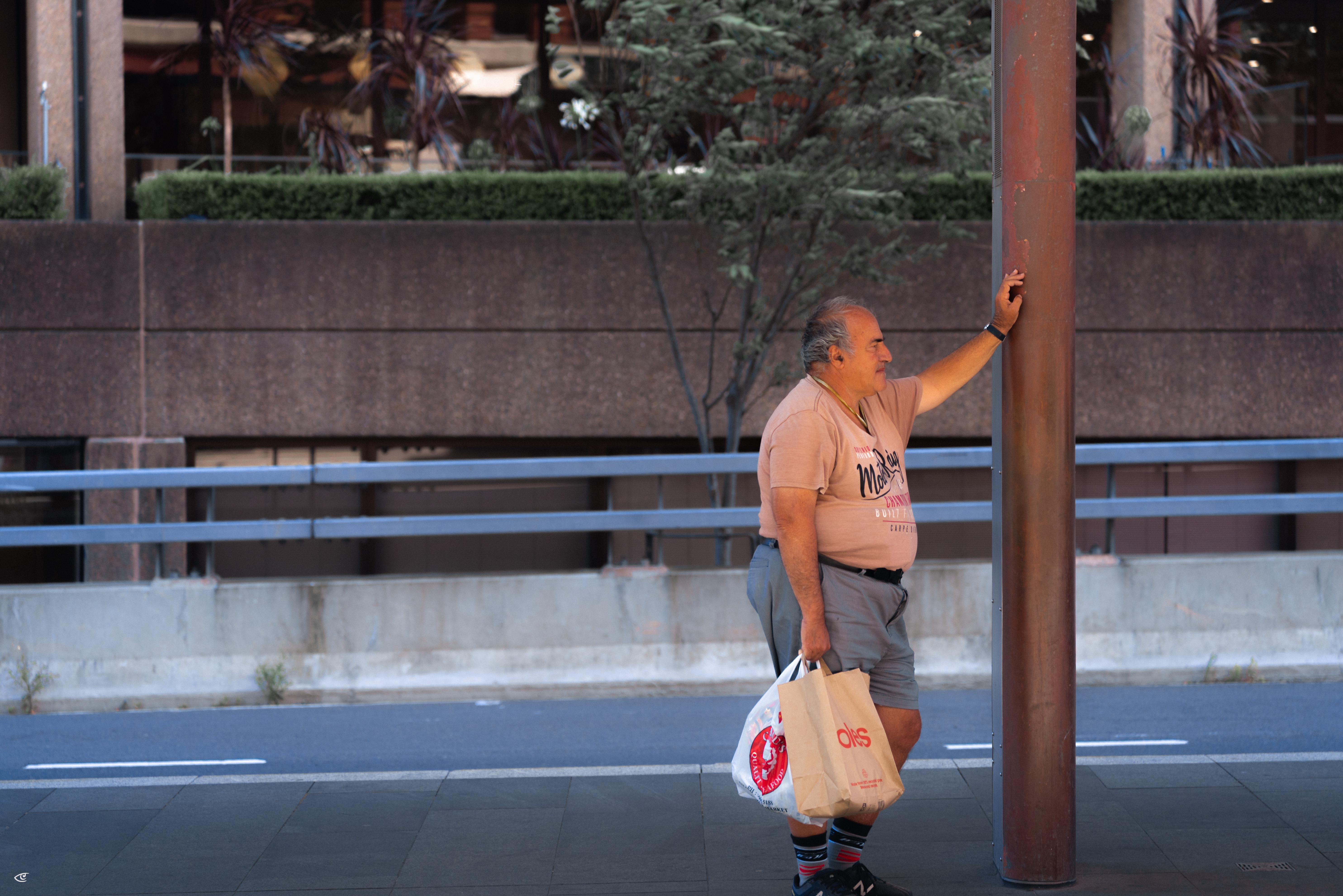 Man standing on a city sidewalk beside a tall brown pole, holding two plastic shopping bags, with a road and concrete wall in the background.