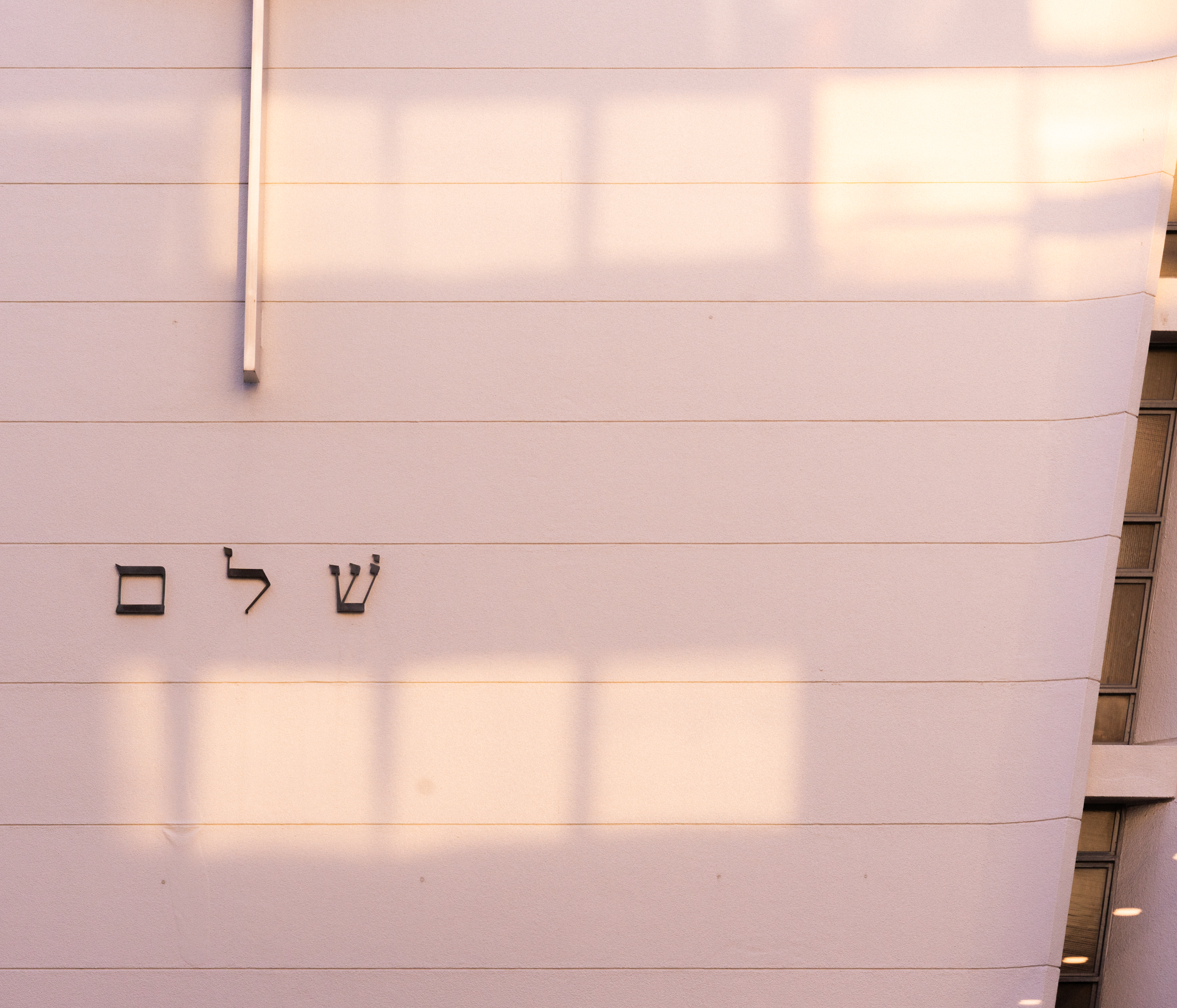 Vertical white wall with horizontal lines and Hebrew letters spelling "שָׁלוֹם" (shalom) near the center. A vertical metal rod is visible on the left side of the image. The wall appears gently curved, with soft shadows creating rectangular patterns.