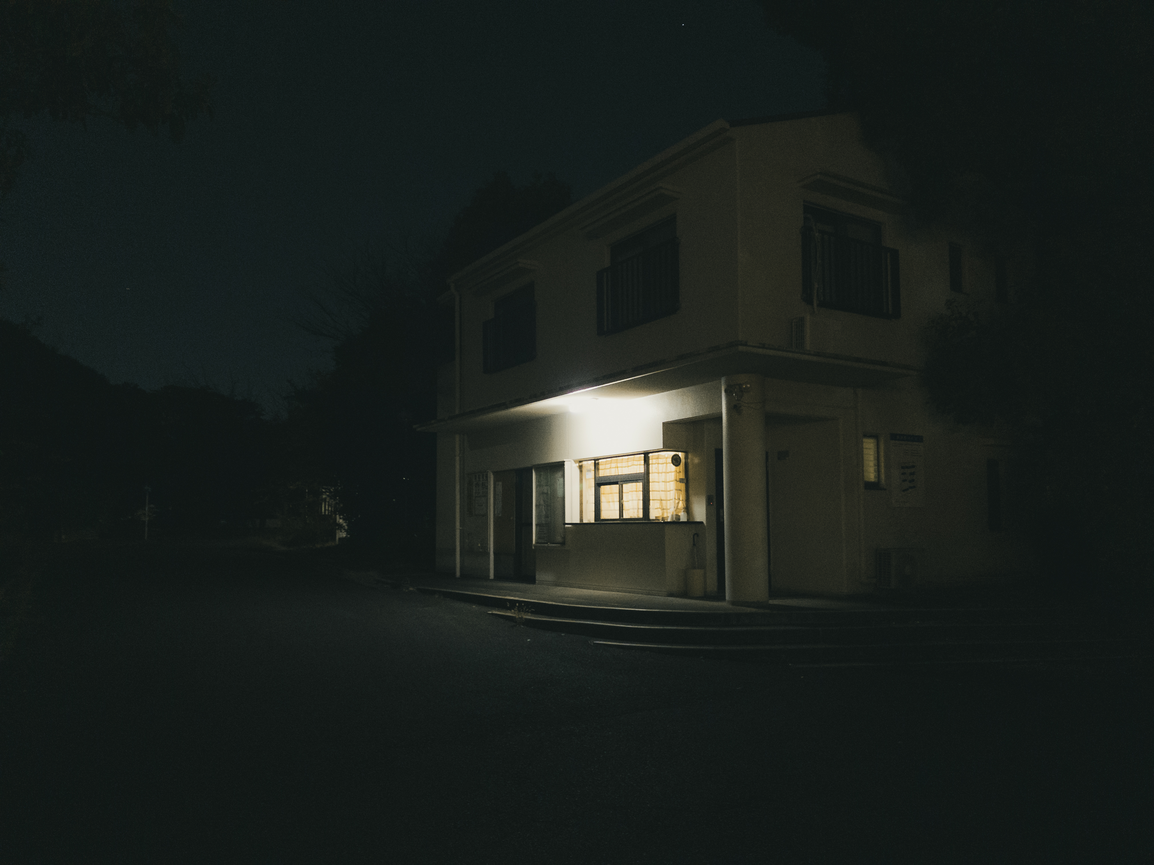 A two-story building with a lighted window on the ground floor. The exterior is dimly lit, with shadows surrounding the structure. A dark, empty street stretches in front, with trees partially visible on the left. The atmosphere appears quiet and nighttime.