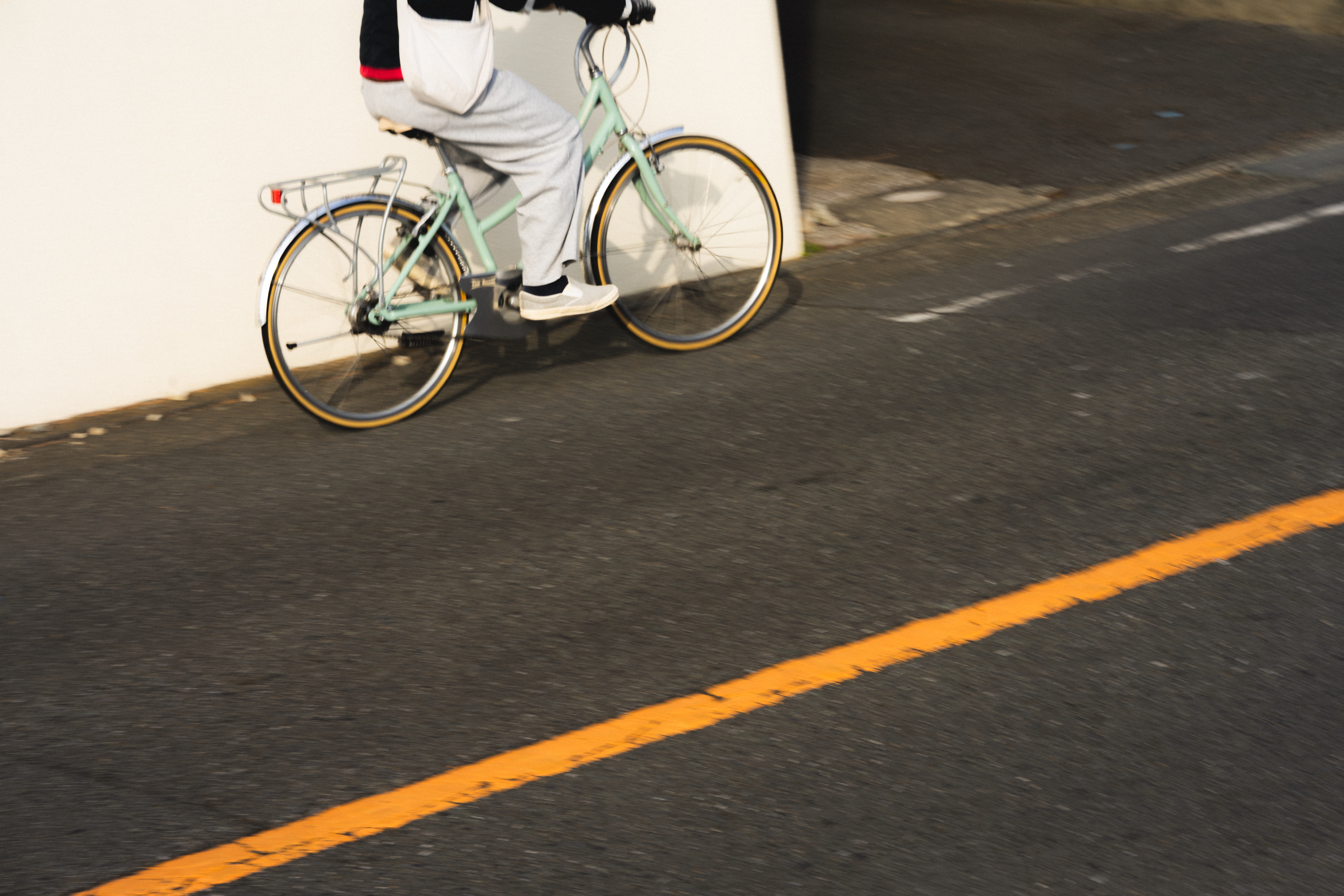 Person riding a light green bicycle with orange tires near a white wall. The road surface is gray with a solid yellow line and faint white dashed lines. The cyclist is wearing gray pants and a dark jacket.