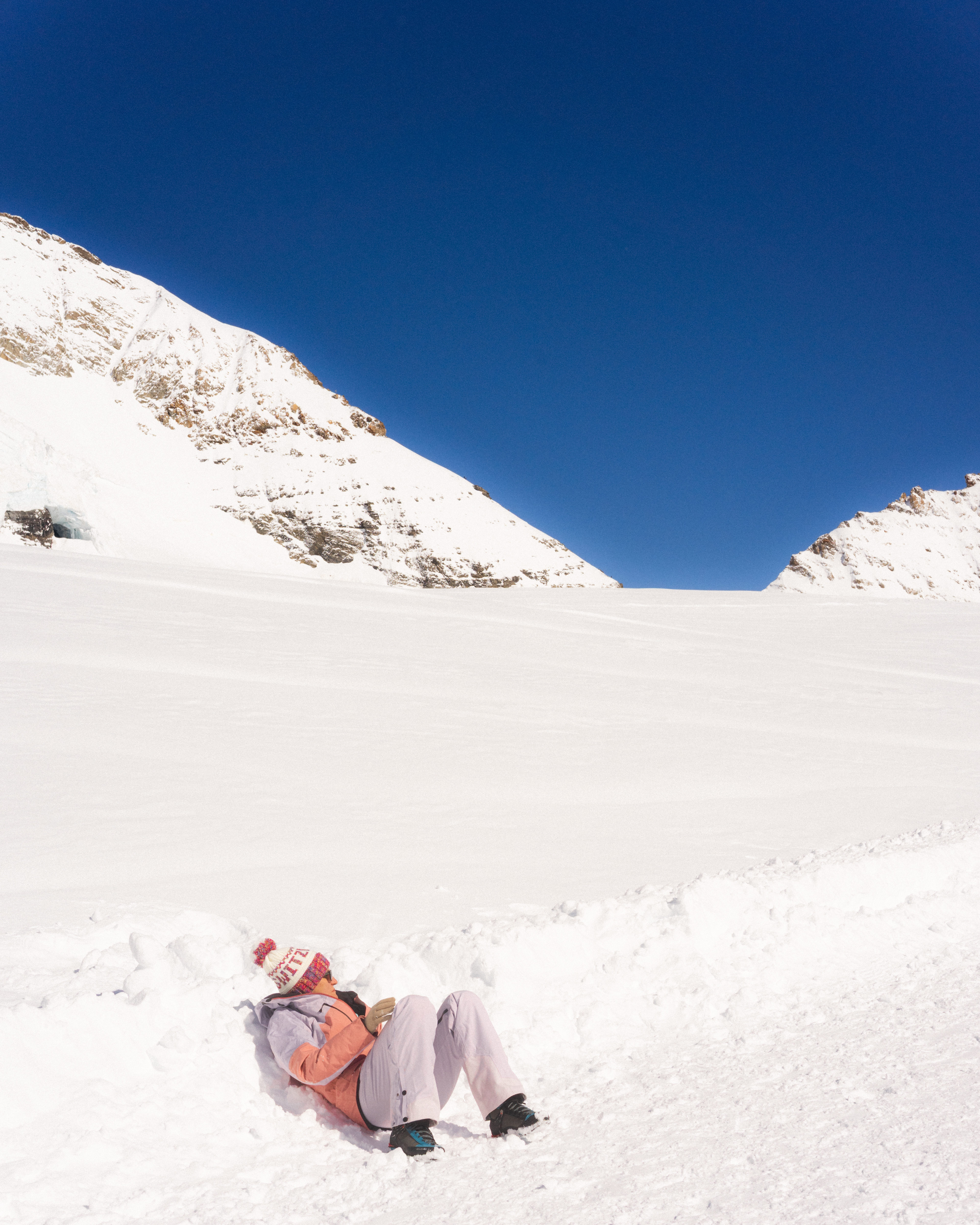 Person in winter clothing sitting on a snow-covered slope, leaning against a snowbank. Wearing a pink jacket and white pants, with a red and white knitted hat. Clear blue sky above and snow-covered mountain peaks in the background.