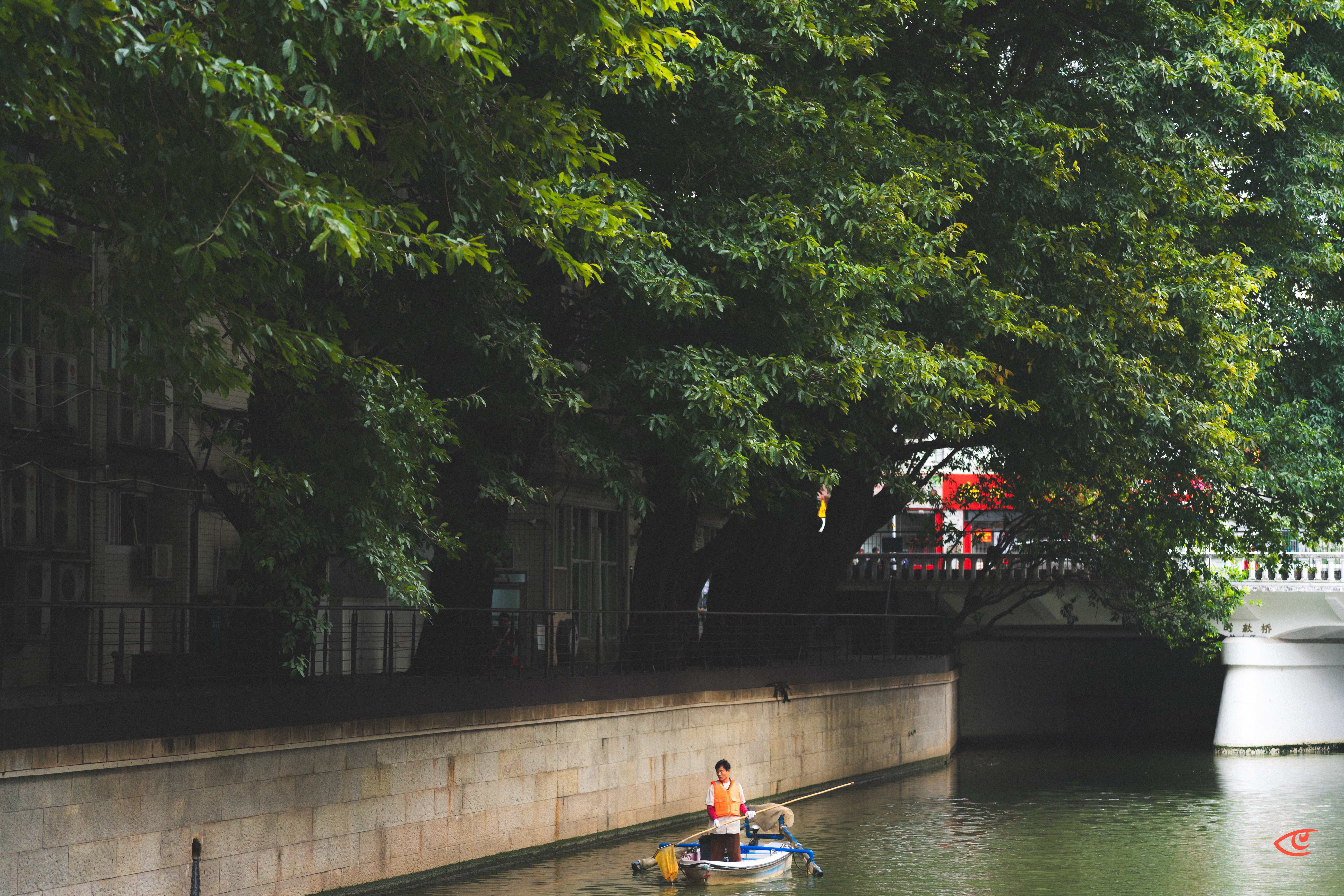 Person in an orange vest standing on a small blue work boat near a stone canal wall, with large overhanging trees shading the water and a bridge in the background.