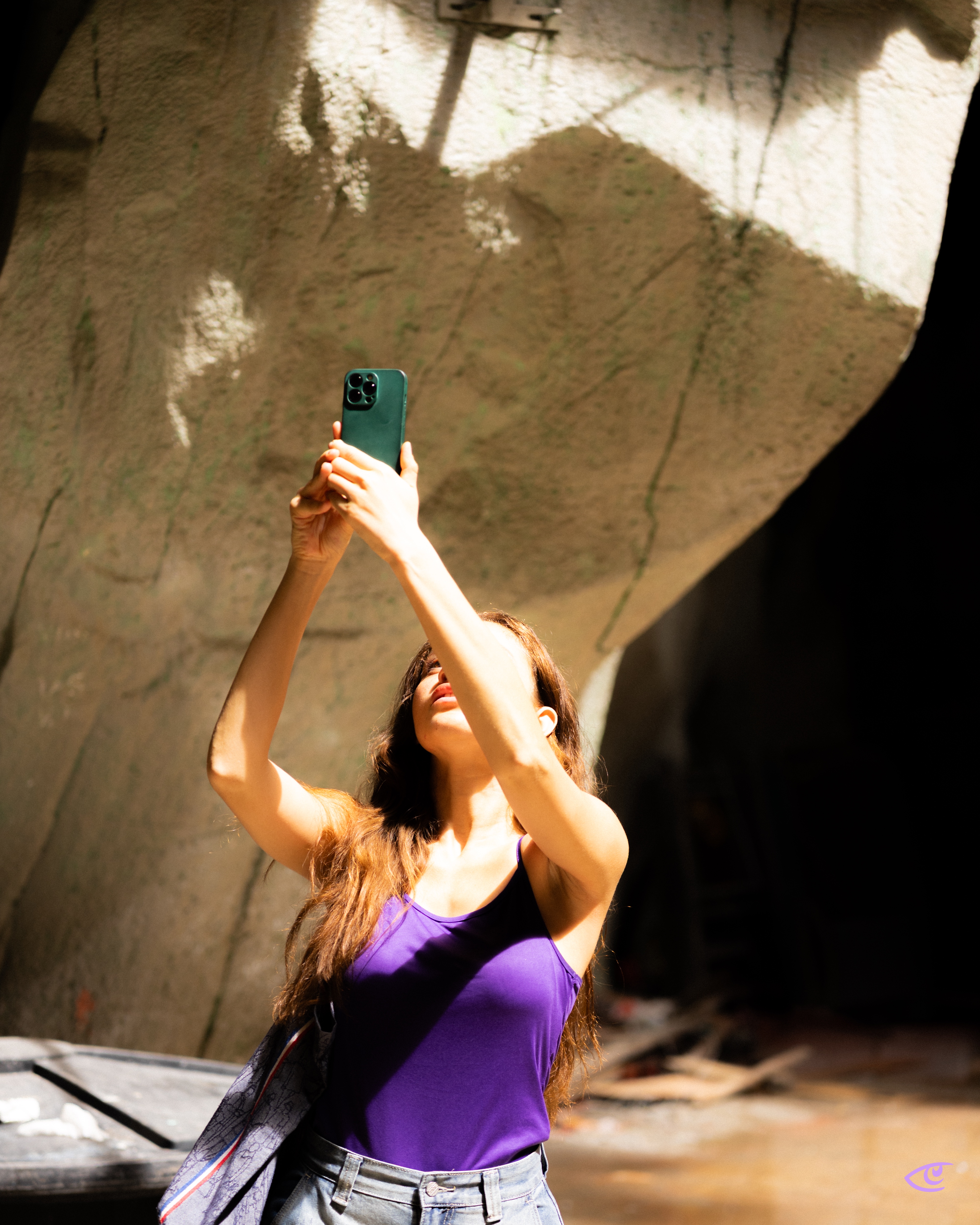 Woman in a purple tank top and jeans holds a green smartphone overhead, looking up as bright sunlight casts large shadows on a rough concrete wall behind her.