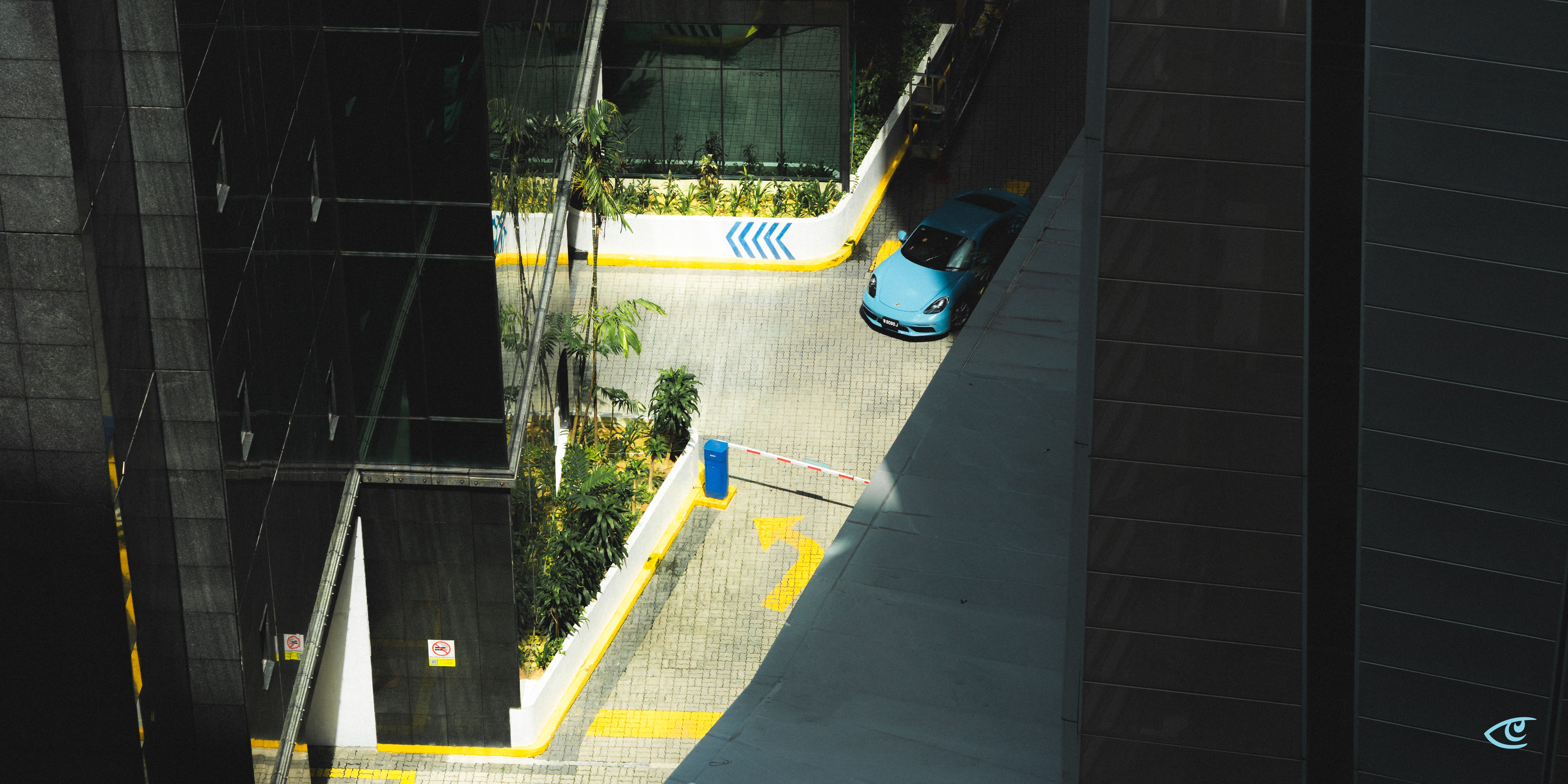 Overhead view of a paved driveway between tall dark buildings, with a blue car near the top right, a red-and-white barrier gate across the lane, and yellow painted arrows and curbs alongside small landscaped plants.