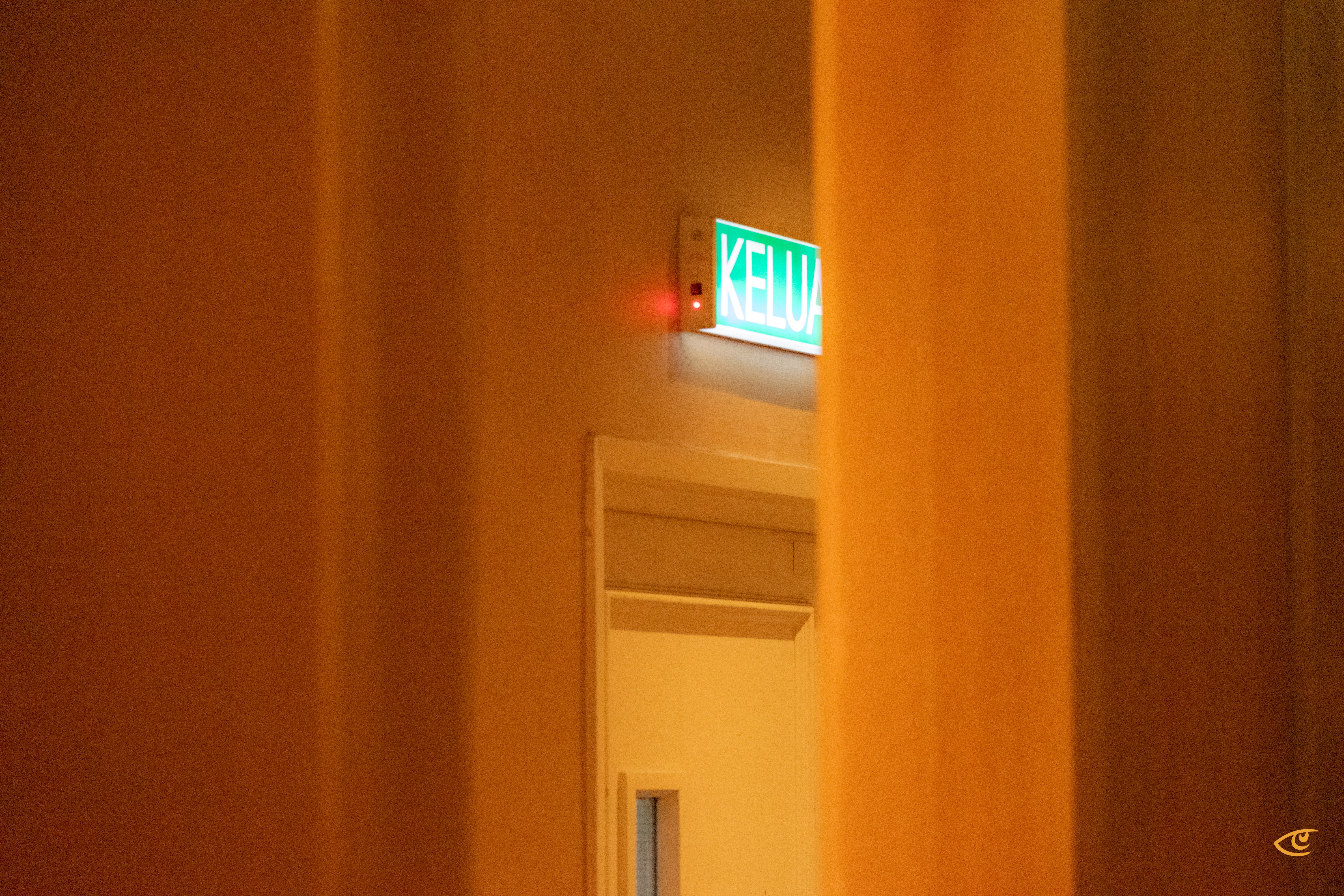 Green illuminated “KELUAR” exit sign above a doorway, viewed through two blurred vertical brown foreground panels that frame the scene.