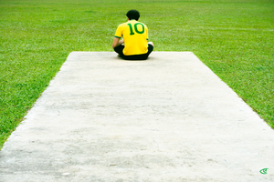 Person sitting cross-legged at the far end of a wide concrete path, viewed from behind, wearing a yellow jersey with a green number 10 and black pants, surrounded by green grass.