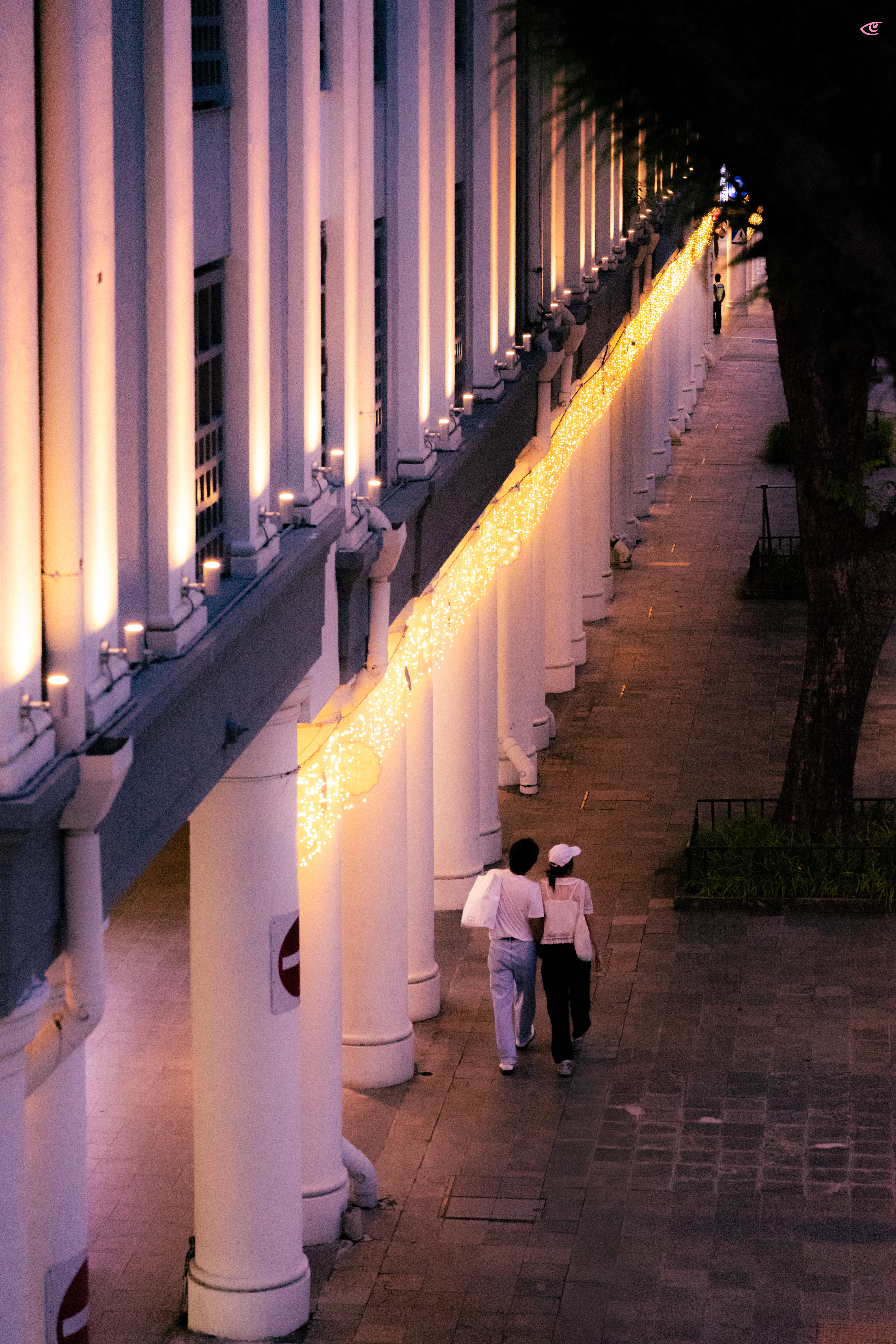 Long colonnade of white pillars along a sidewalk at dusk, lit by a continuous string of warm fairy lights; two people walk away in the foreground while trees line the right side.