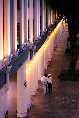 Long colonnade of white pillars along a sidewalk at dusk, lit by a continuous string of warm fairy lights; two people walk away in the foreground while trees line the right side.