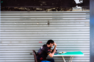 Man sitting alone at a green table in front of a closed corrugated metal shutter, leaning forward with his head resting on his hand; a plate of food and a drink sit on the table.