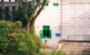 White building exterior with bright green shuttered windows, one open above a driveway barrier; a blue “Pedestrian Walkway” sign on the wall, trees and dense shrubs in the foreground, and a paved courtyard with a low fence on the right.
