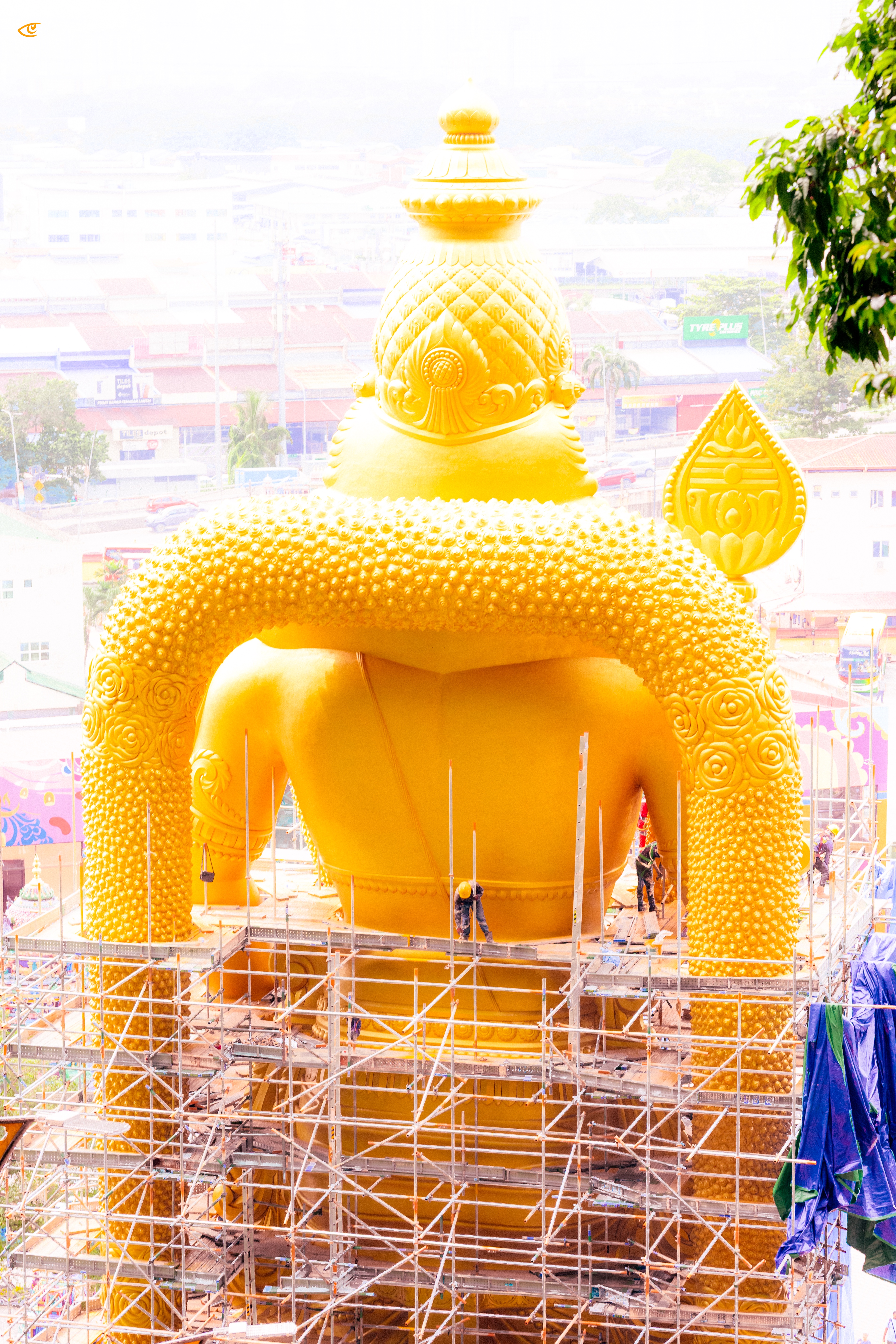 Rear view of a large golden Buddha statue with an ornate curled-hair texture and pointed headpiece, surrounded by metal scaffolding and a few workers; a hazy cityscape and tree branches appear in the background.